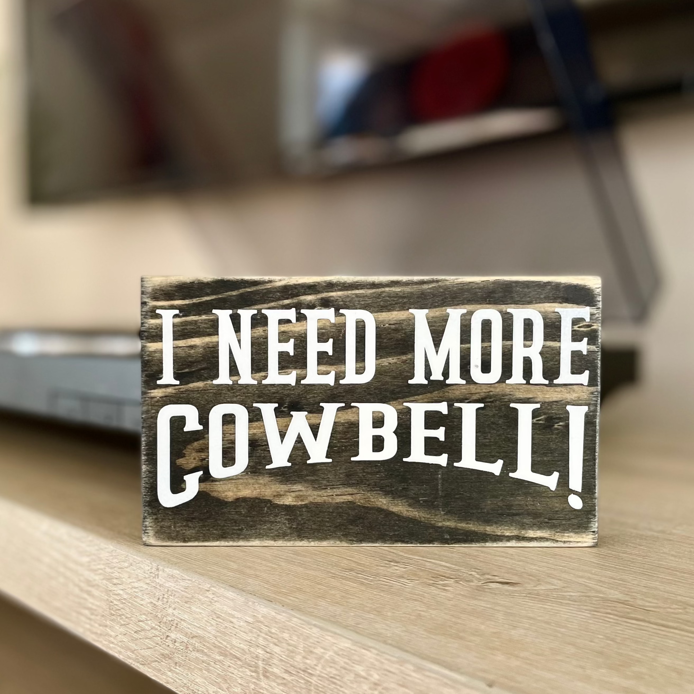 A small, black, wood sign sits on a shelf with a record player blurred behind it.