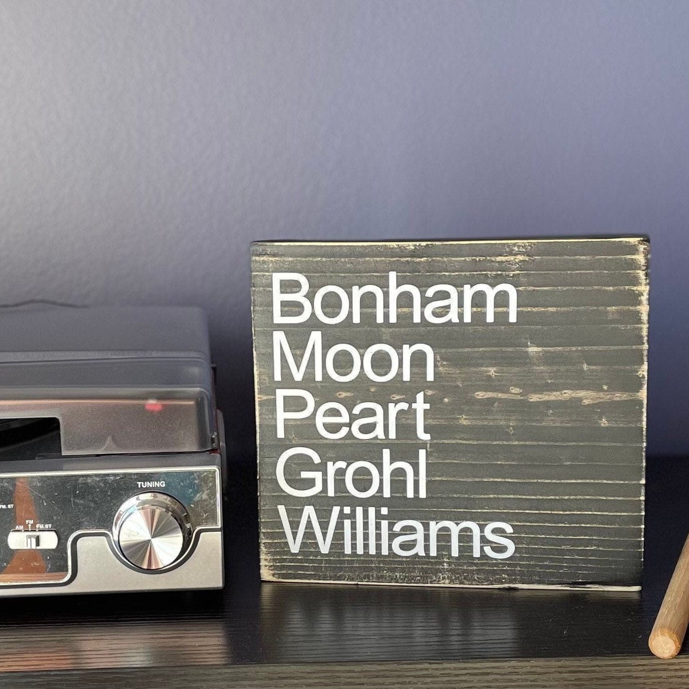 A black, wood sign sits on a table with drumsticks & a record player.