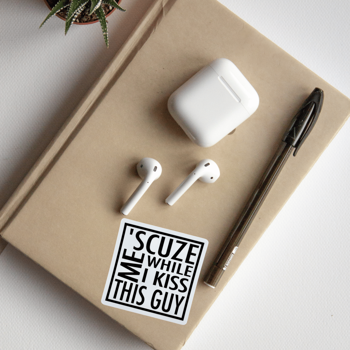 White wireless earbuds and charging case on a beige surface with a pen and 'SCUZE ME WHILE I KISS THIS GUY' sticker.