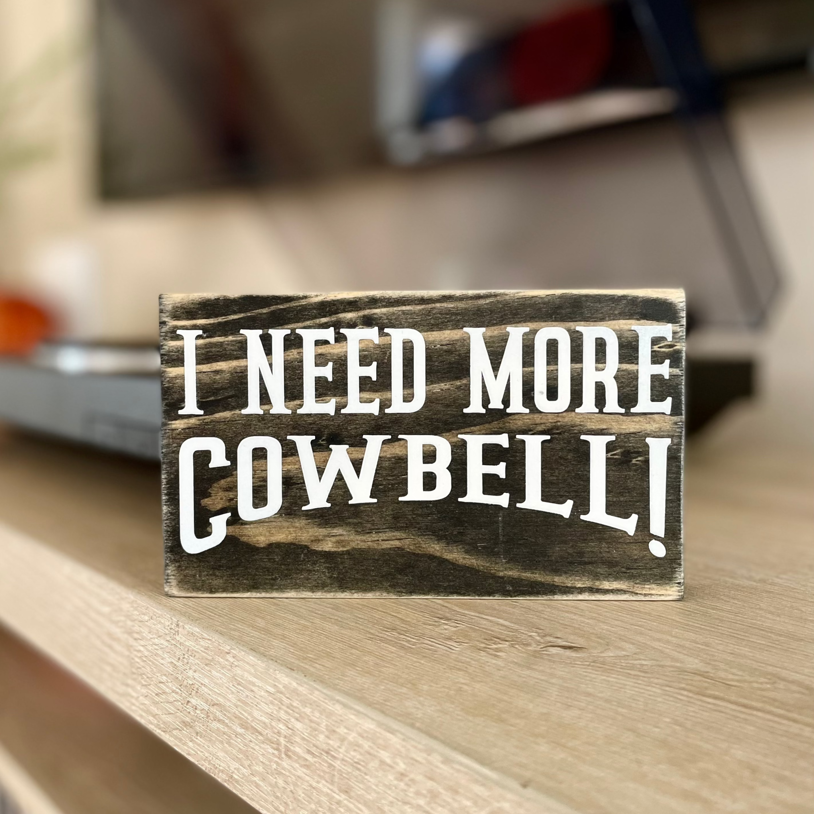 A small, black, wood sign sits on a shelf with a record player blurred in the background.