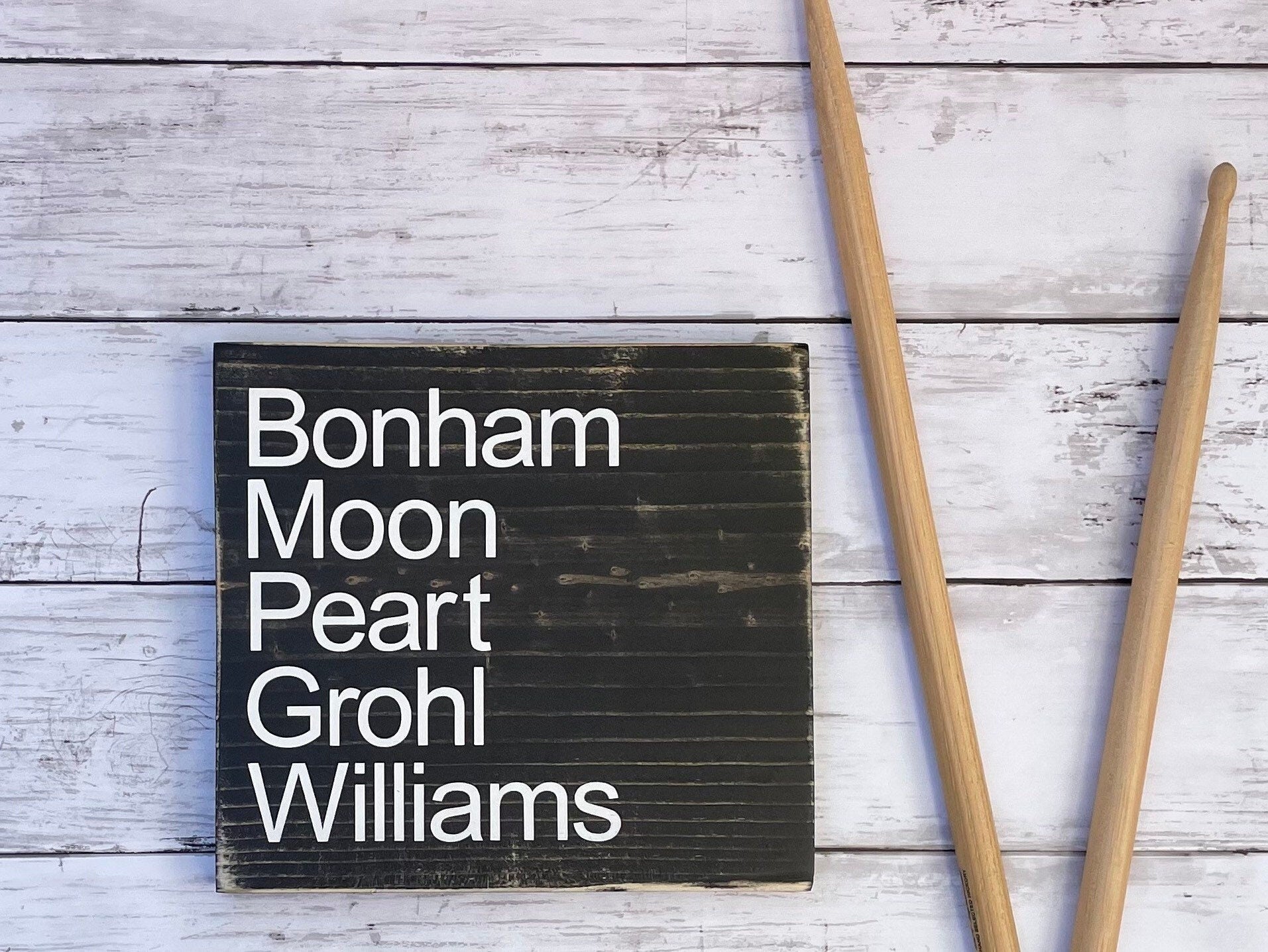 A black, wood sign sits on a. table with drumsticks