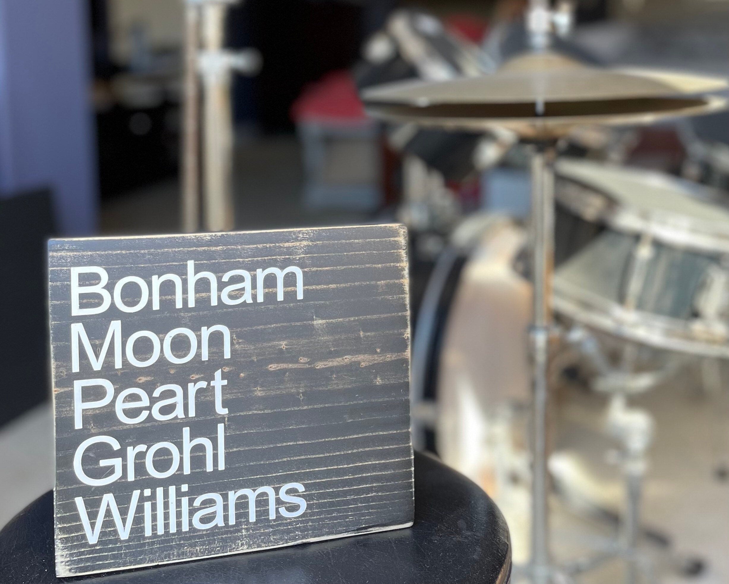 A black, wood sign sits on a drum throne with a drum kit in the background.