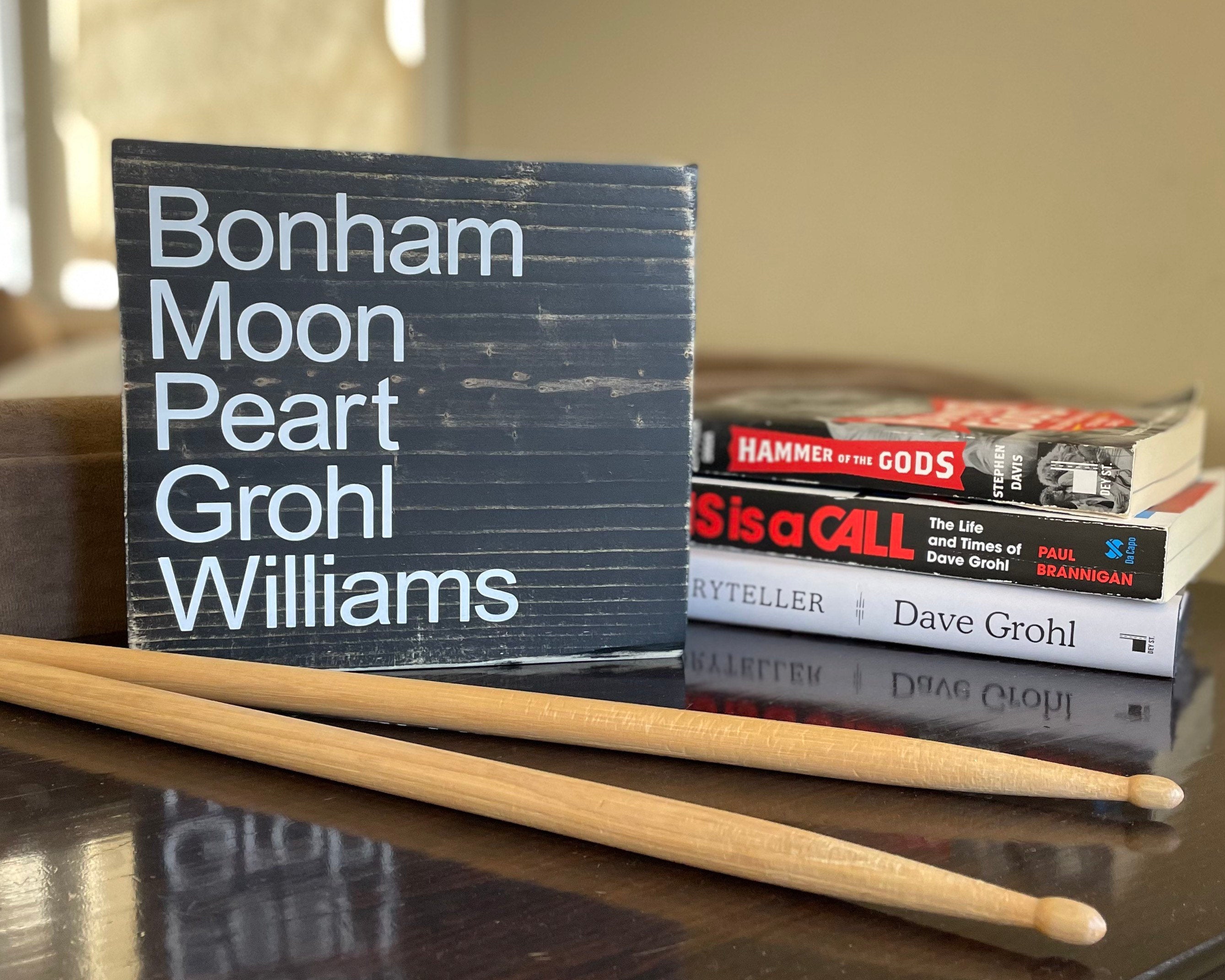A black, wood sign sits on a. table with drumsticks & music-themed books