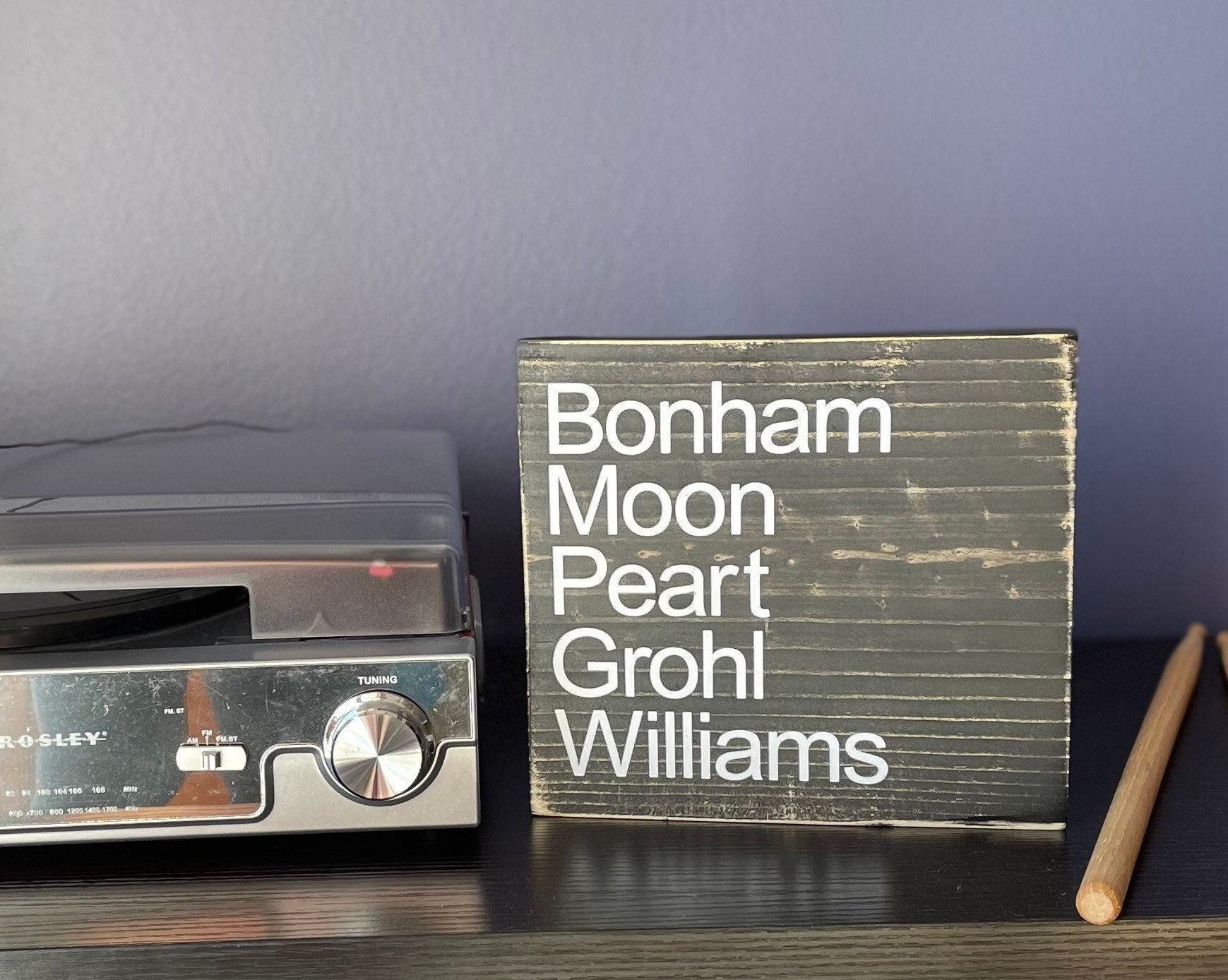 a square wood sign stained black with white, left justified, lettering that reads "Bonham Moon Peart Grohl Williams". The names are listed top to bottom.