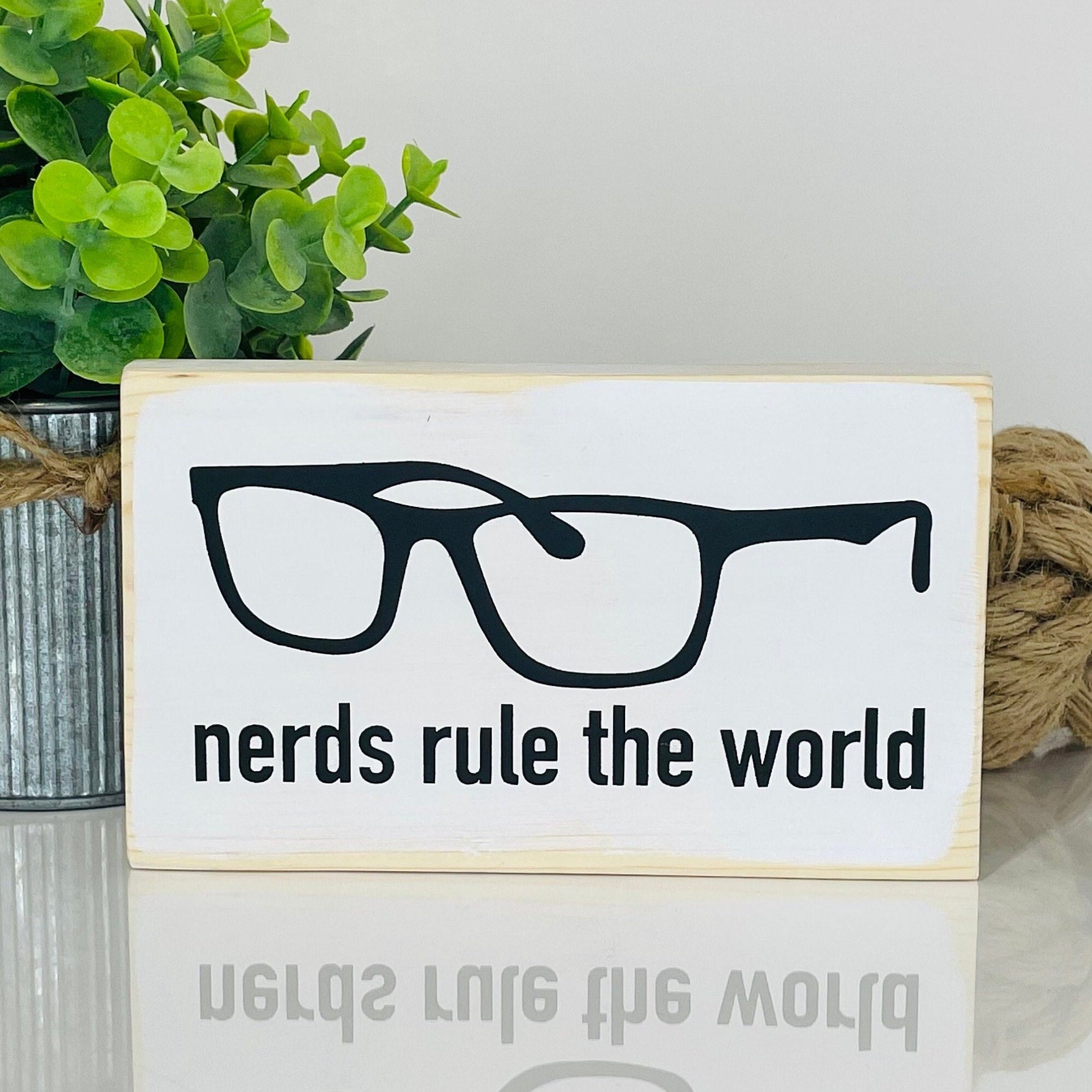 small wood rectangular sign painted white with black lettering in all lowercase that reads "nerds rule the world". Words are at the bottom of the sign. Above the words is the outline of a black pair of nerdy-style glasses