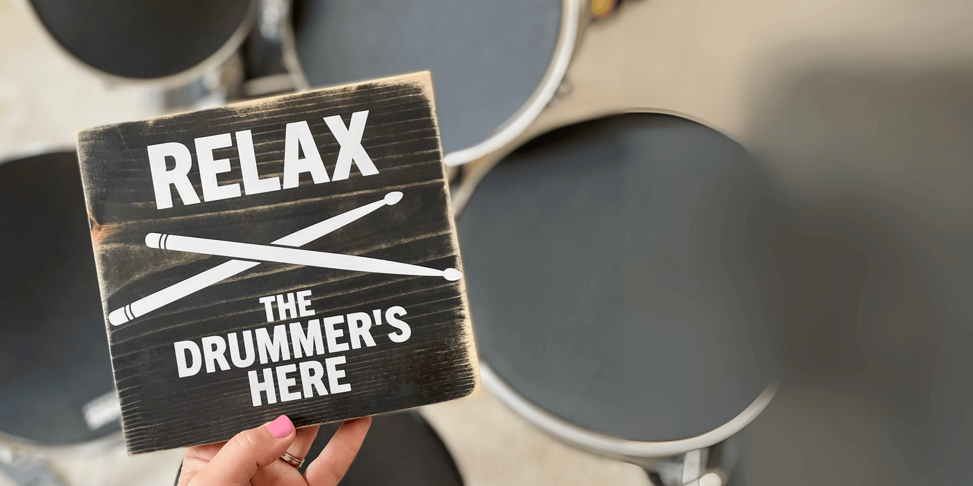A hand is holding a black, wood sign that says Relax the drummer's here. A drum kit is blurred in the background
