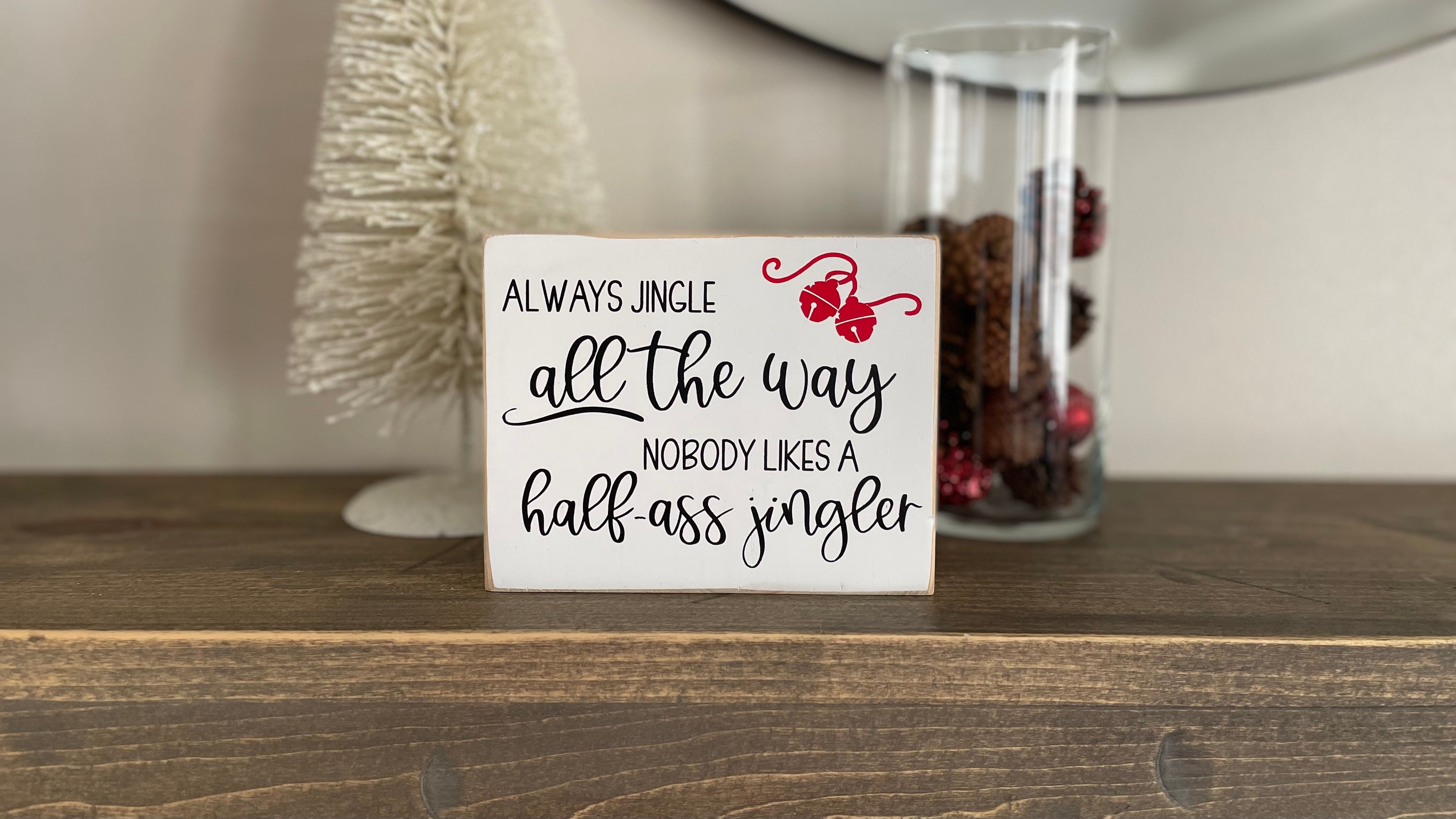 small wood sign painted white with black text that reads "always jingle all the way nobody likes a half ass jingler". Two red bells on a ribbon are in the top right corner