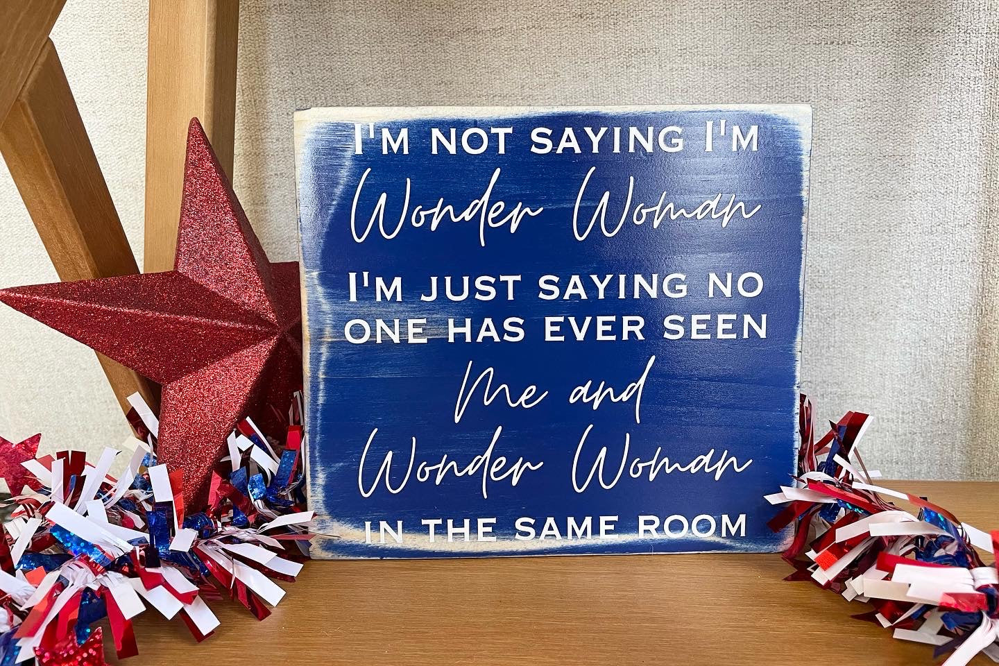 medium sized square wood sign painted blue with white lettering that reads, " I'm not saying I'm Wonder Woman, I'm just saying no one has ever seen me and Wonder Woman in the same room". All text is in all caps except for "Wonder Woman" and "Me and Wonder Woman". Text is centered, takes up the entire area and is on 7 lines. Glossy finish.