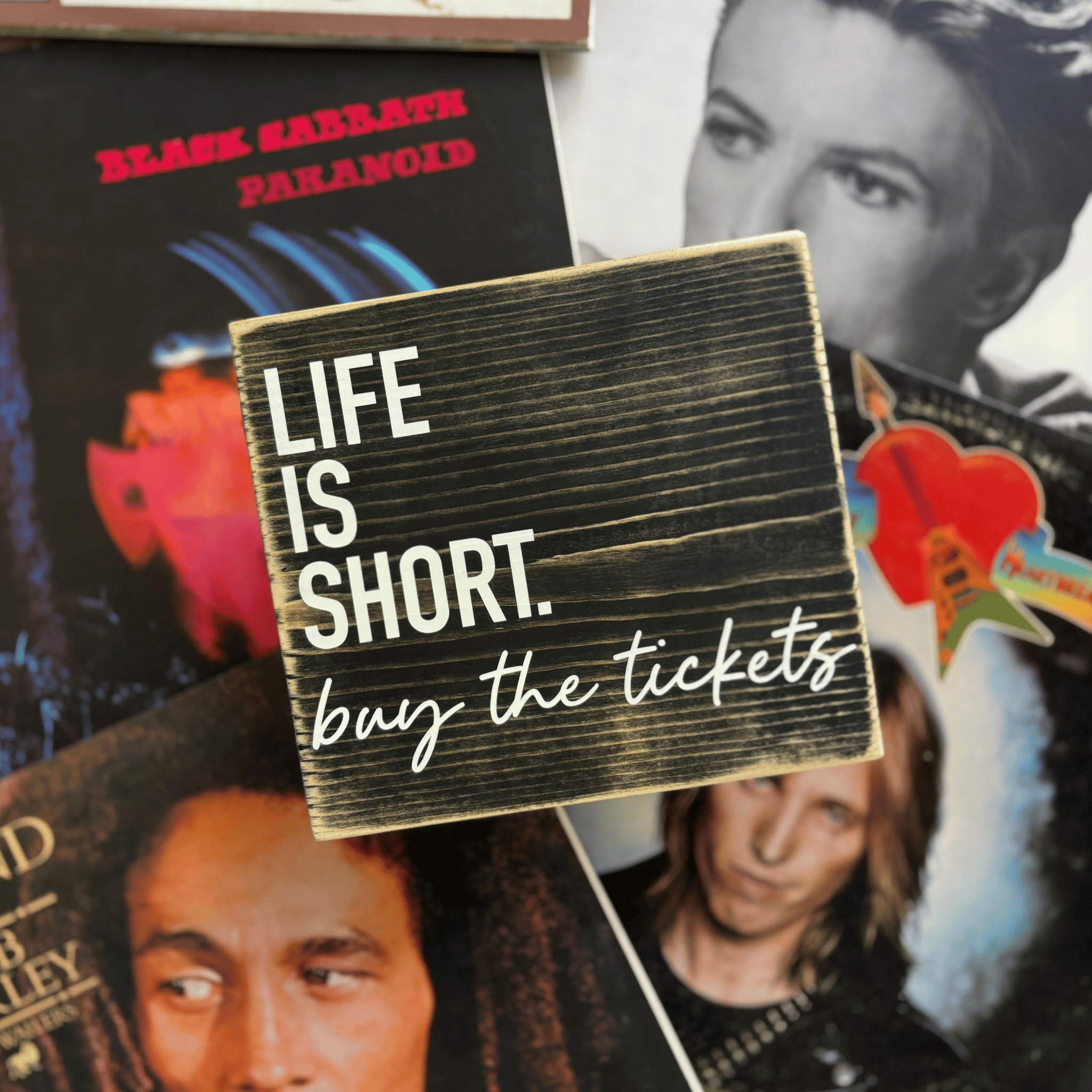 Small wooden sign with 'Life is Short, buy the tickets' text on a background of album covers and photos.