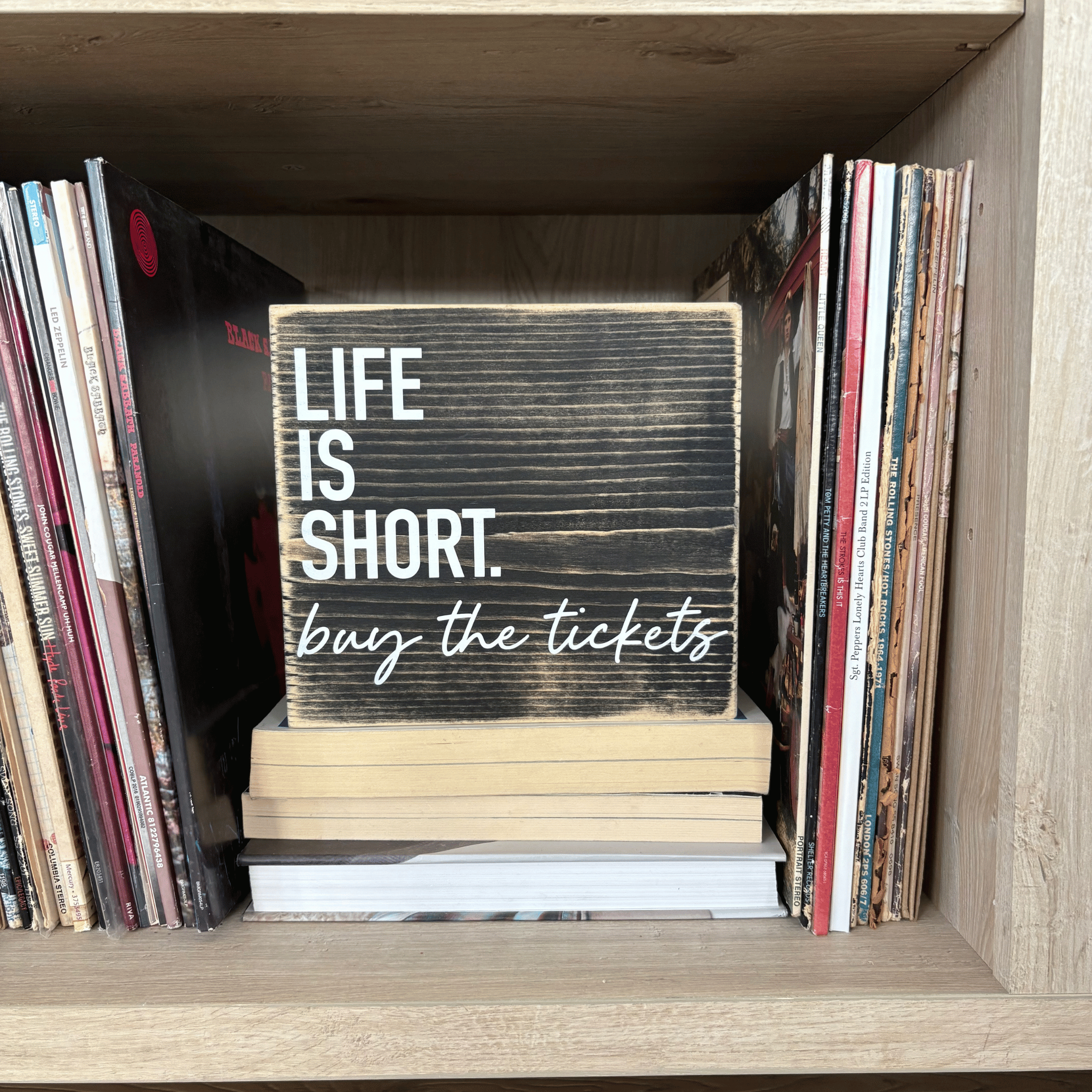 Wooden sign with 'Life is Short, buy the tickets' text stacked on books in front of vinyl records