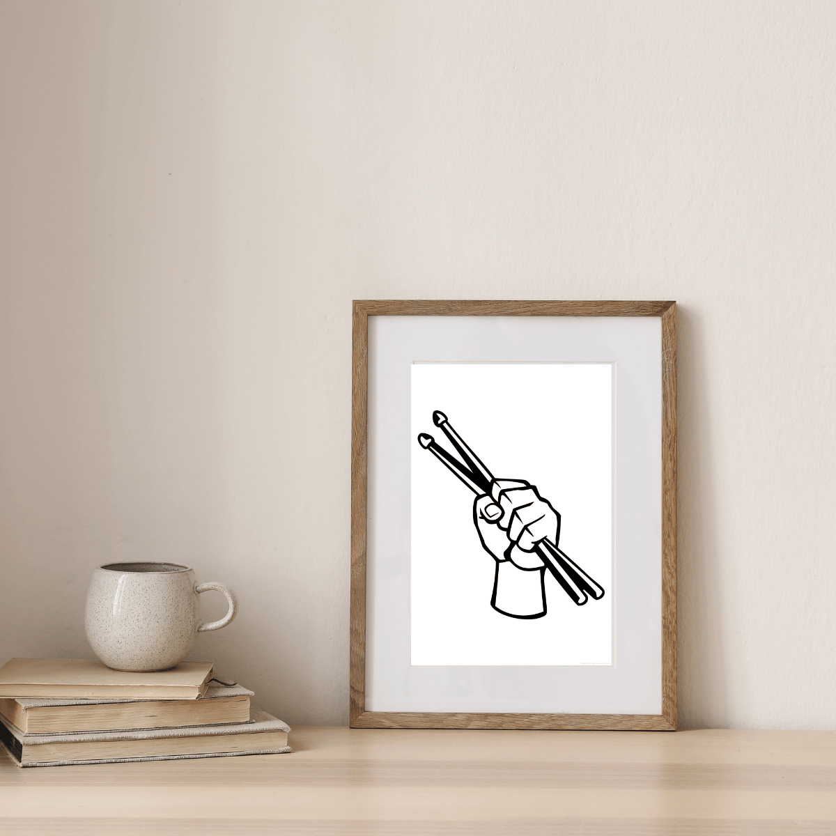 A framed minimalist art print featuring a black line drawing of a hand holding drumsticks, displayed on a light wood table next to a ceramic mug and books against a neutral beige wall.