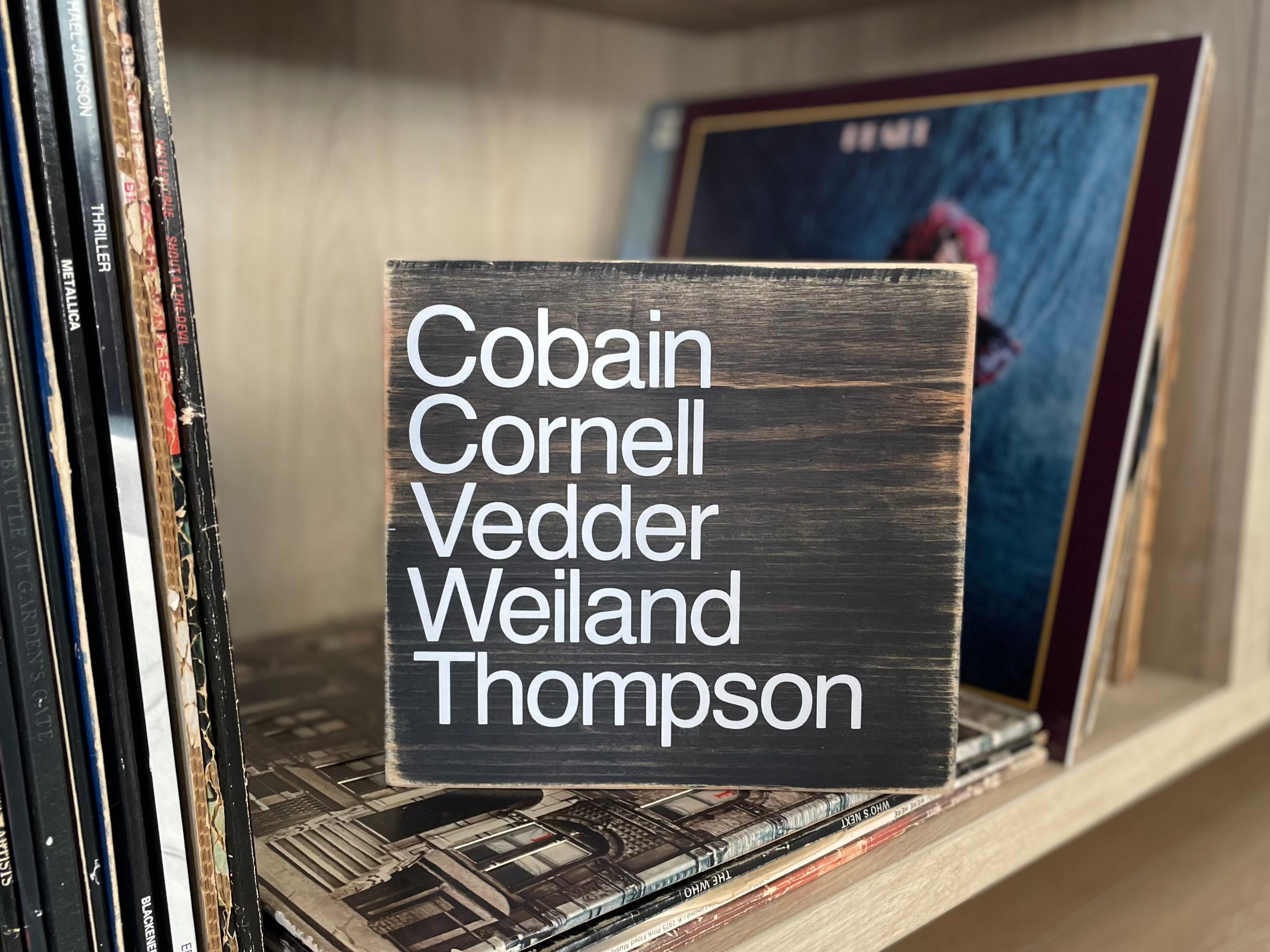 A square, black, wood sign sits on a shelf stacked with vinyl records.