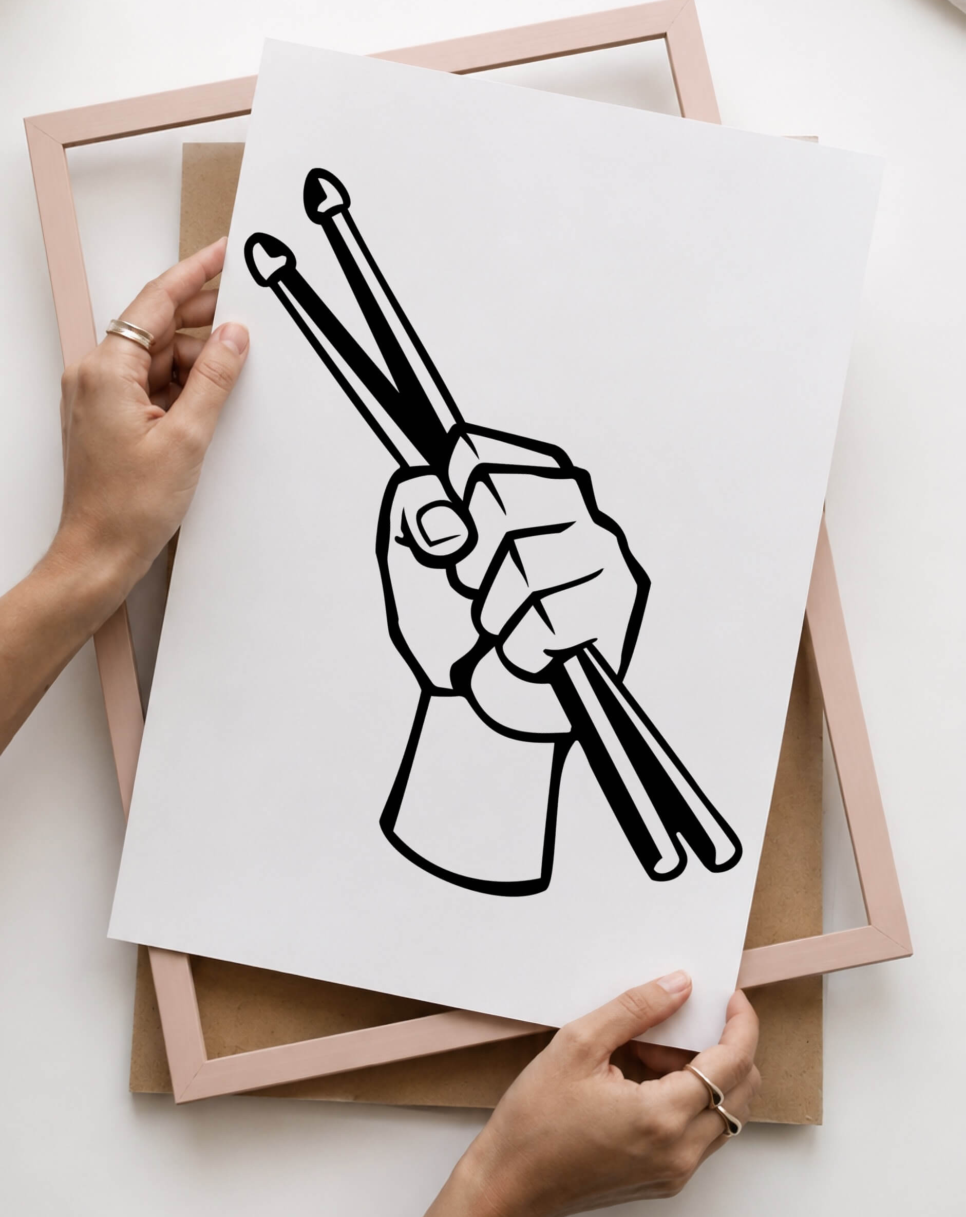 A person holds a white art print preparing to put it in the disassembled frame on their work surface. The print features a black line art illustration of a hand holding drumsticks. 