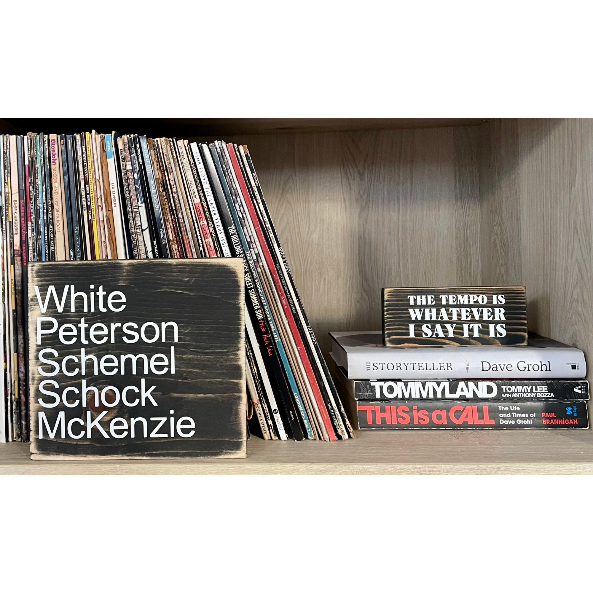 Two black, wood signs sit in a bookcase with vinyl records. The larger sign reads in white "White Peterson Schemel Schock McKenzie" The smaller sign reads, "THE TEMPO IS WHATEVER I SAY IT IS"