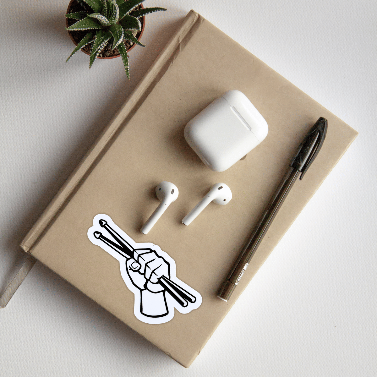 White airpods, airpod case, pen, and sticker on a beige notebook with a plant in the corner. A sticker of drumsticks held in a fist is on the notebook.