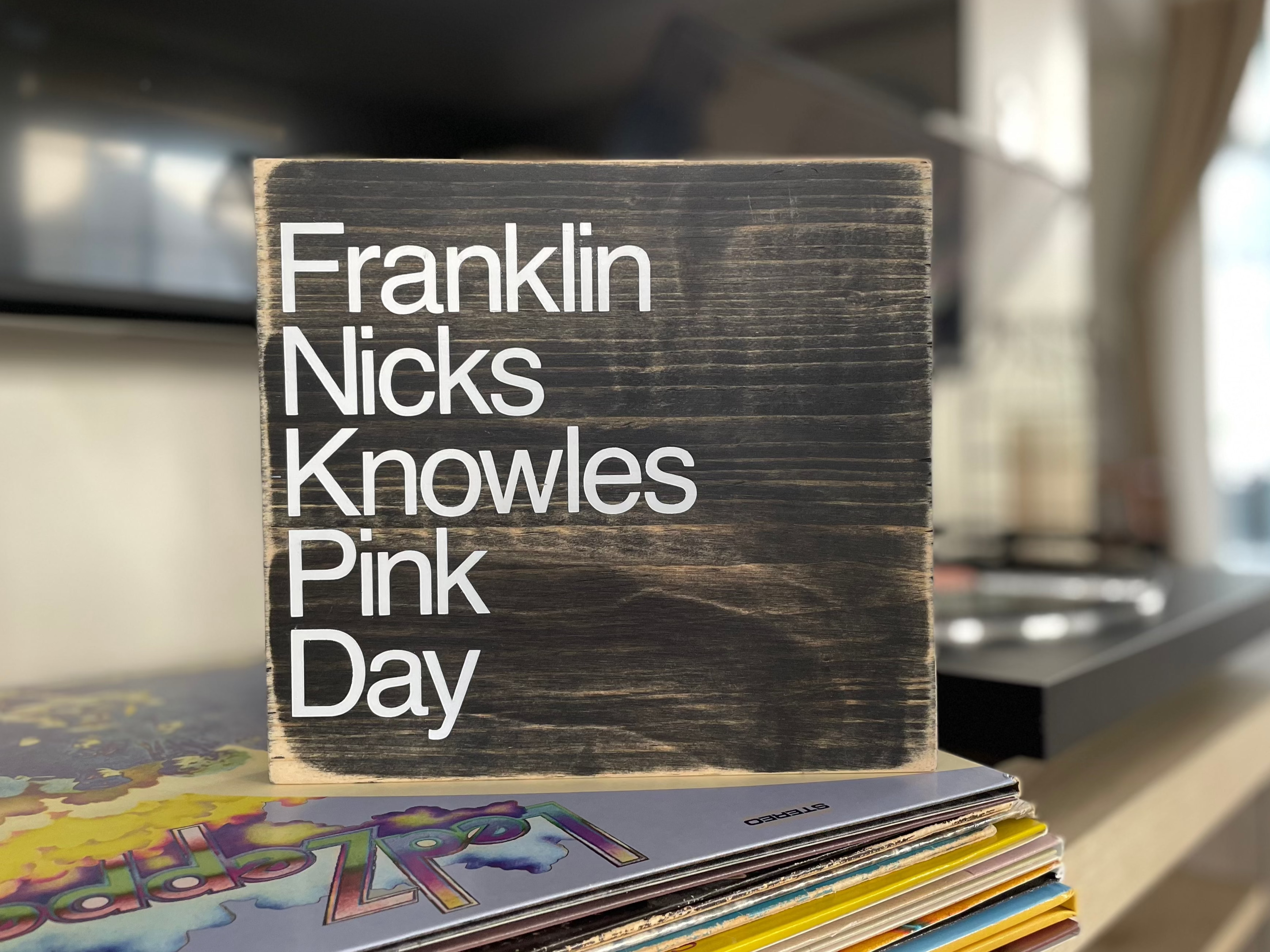 A square, black, wood sign sits on a table next to a record player.