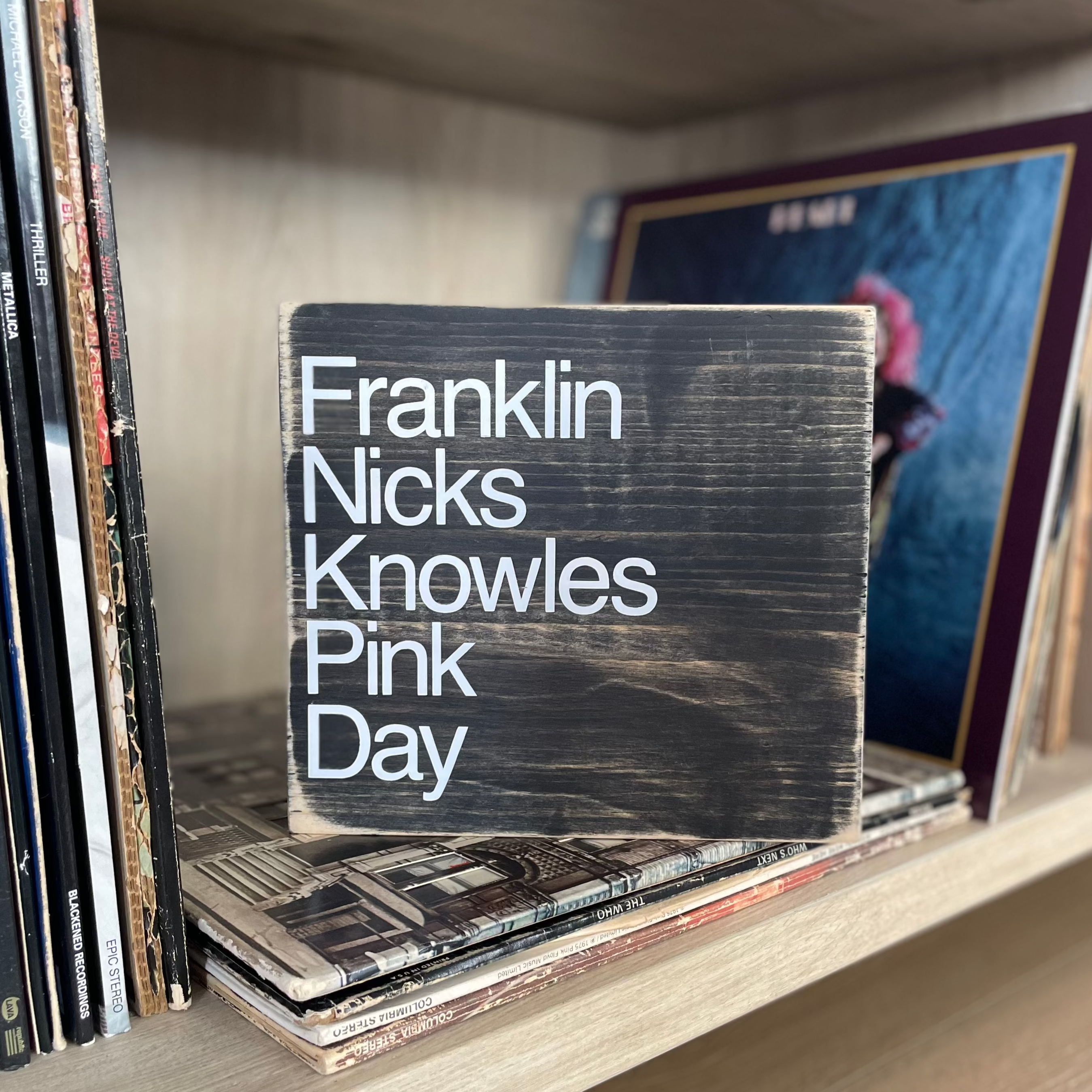 A square, black, wood sign sits on a shelf stacked with vinyl records.