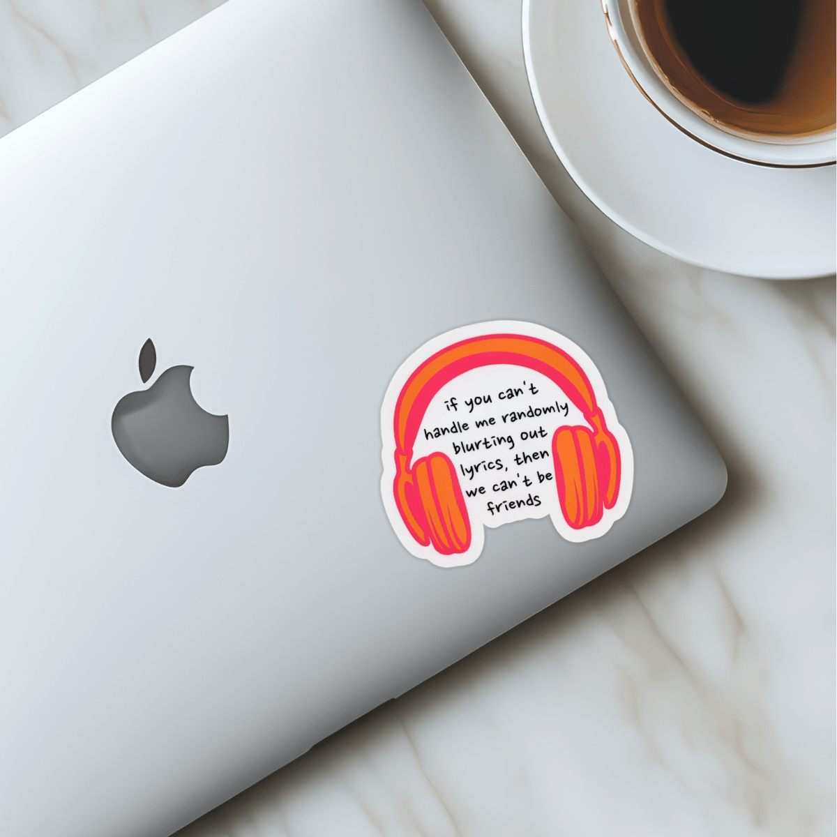 Laptop with a sticker of headphones and text next to a cup of coffee on a marble surface