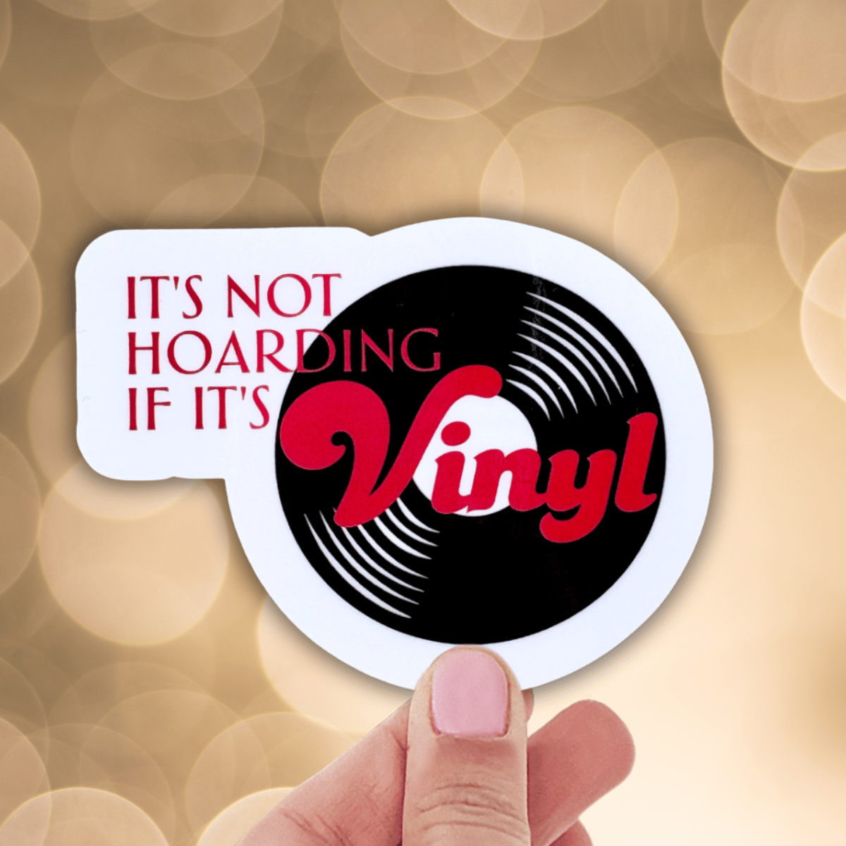 Sticker with vinyl record design and text 'It's not hoarding if it's vinyl' held by a hand against a blurred background.