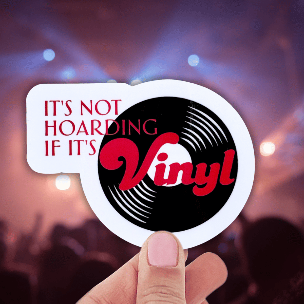 Sticker with vinyl record design and text 'It's not hoarding if it's vinyl' held by a hand against a blurred concert background.
