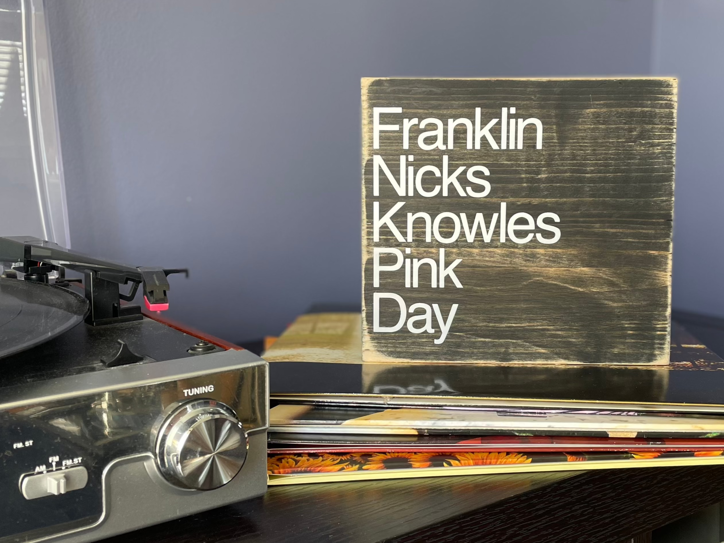 A square, black, wood sign sits on a table next to a record player.