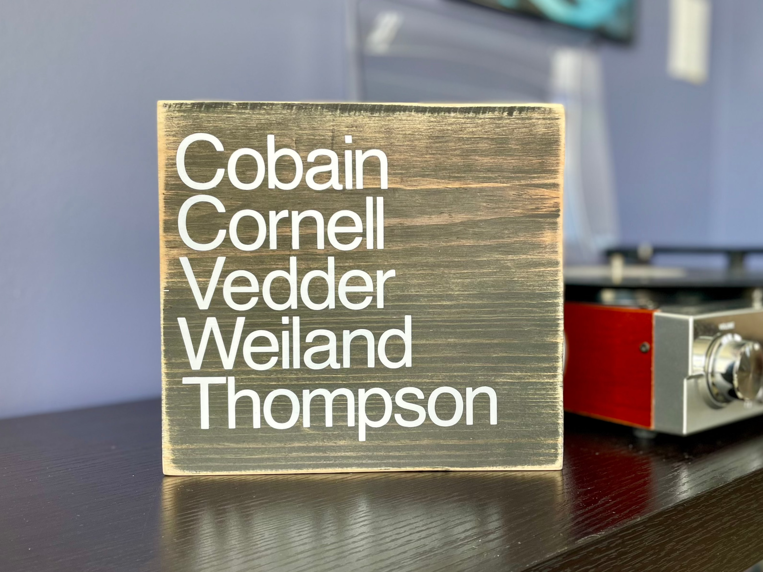 A square, black, wood sign sits on a table next to a record player.