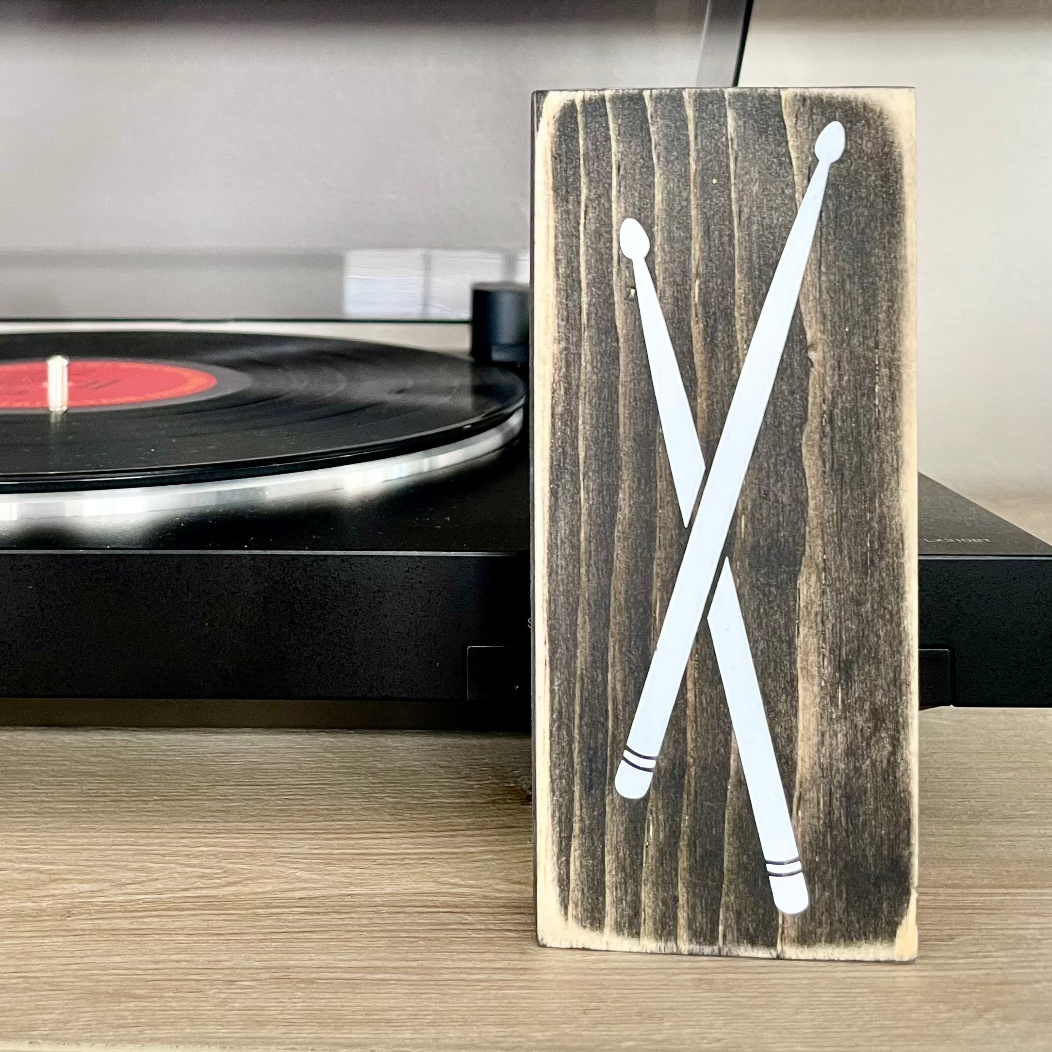 A small, black, wood sign with drumsticks painted on it, sits vertically on a table in front of a record player. 