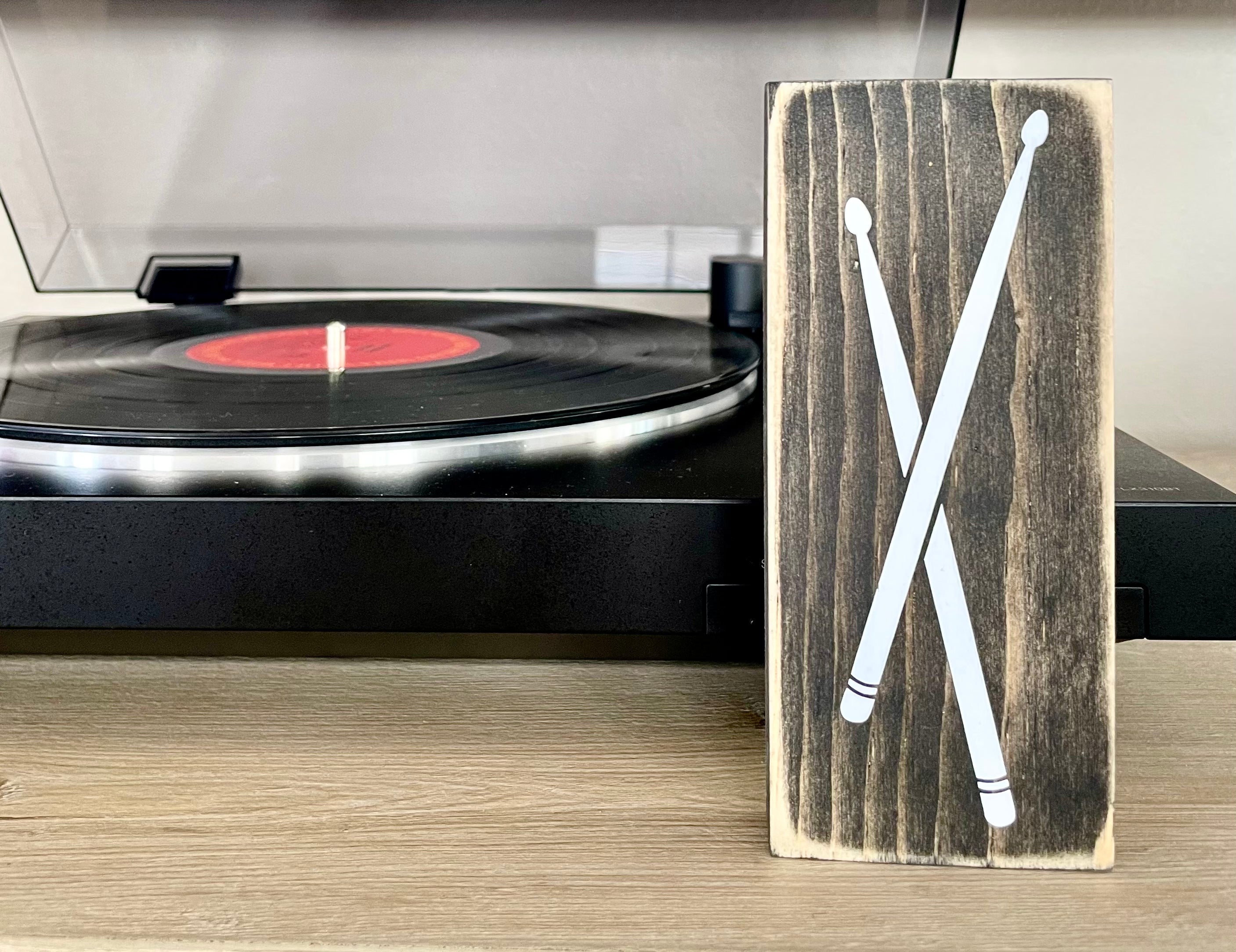 A small, black, wood sign with drumsticks painted on it, sits vertically on a table in front of a record player. 