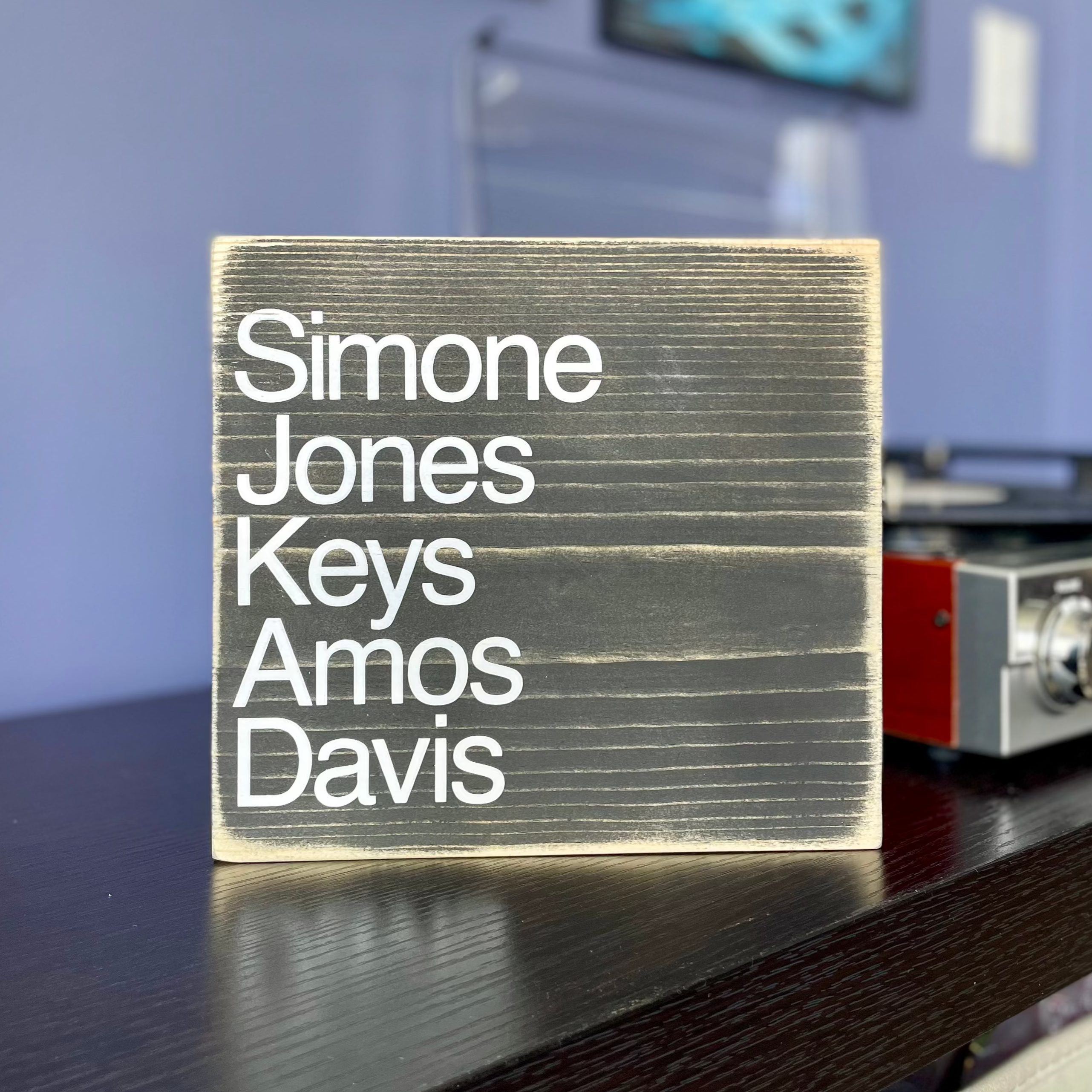 A square, black, wood sign sits on a table next to a record player