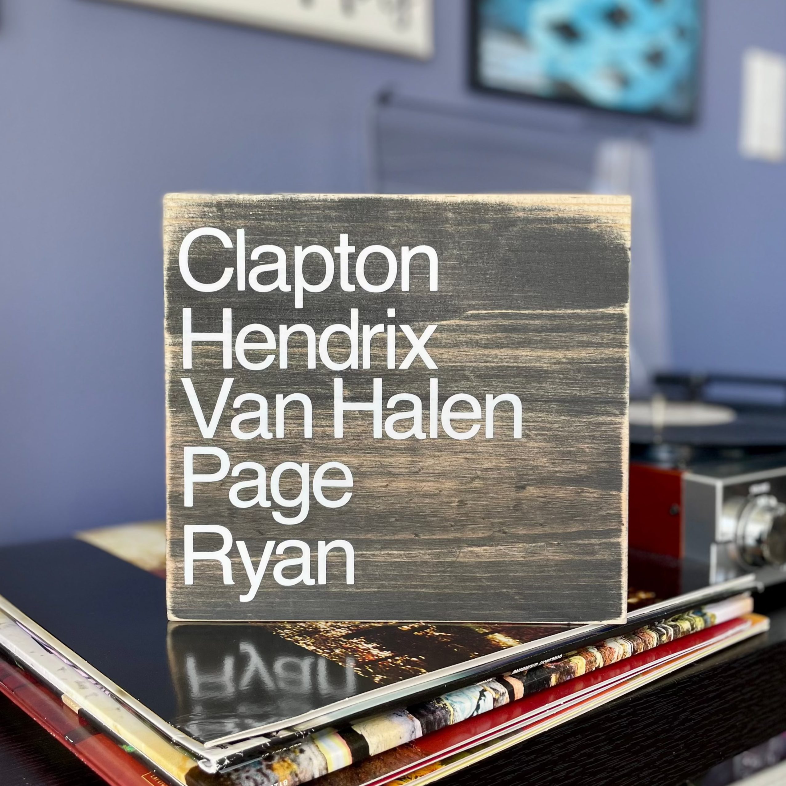 A square, black, wood sign sits on a stack of vinyl records next to a record player.