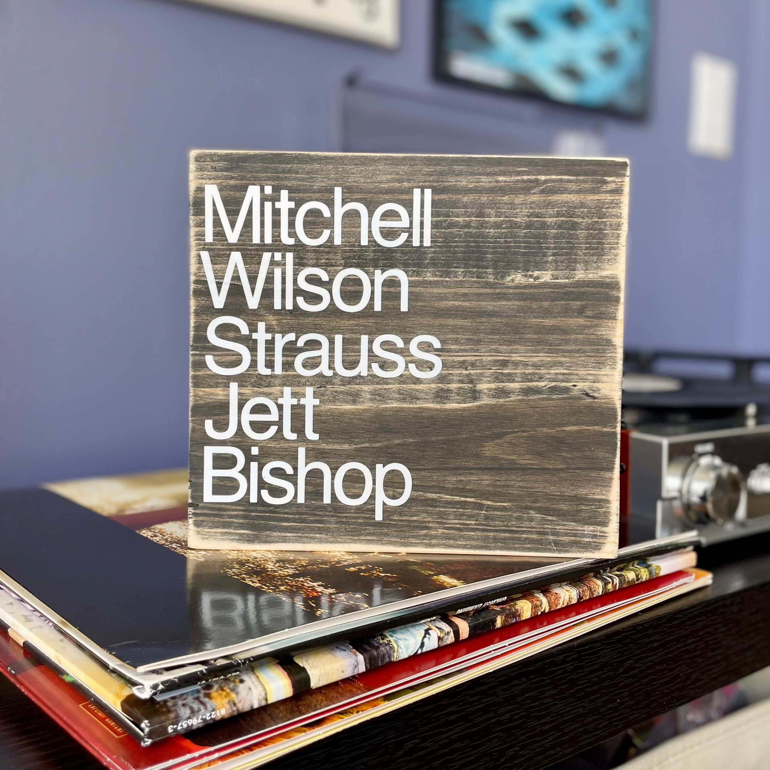 A square, black, wood sign sits on a stack of vinyl records next to a record player.