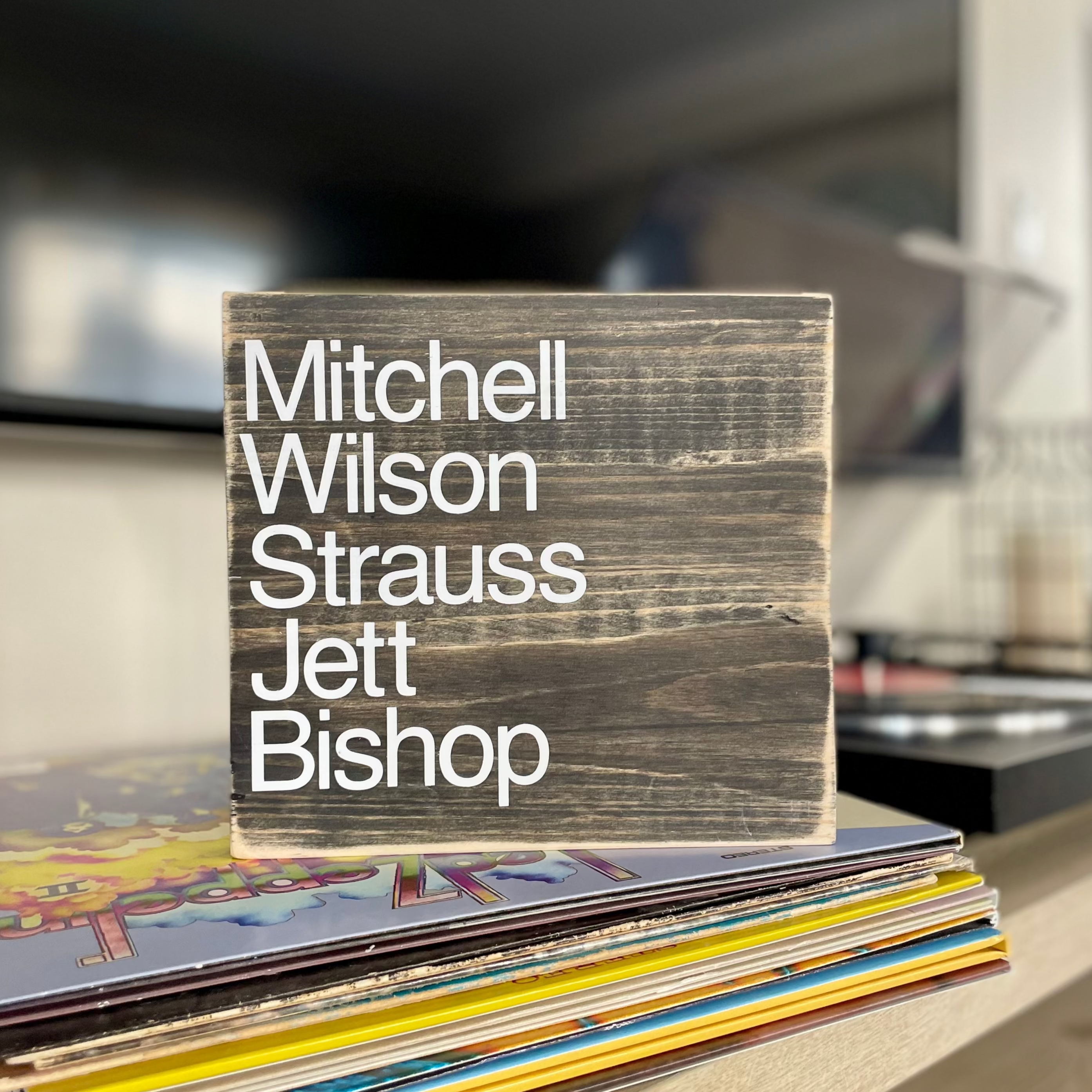 A square, black, wood sign sits on a stack of vinyl records next to a record player.