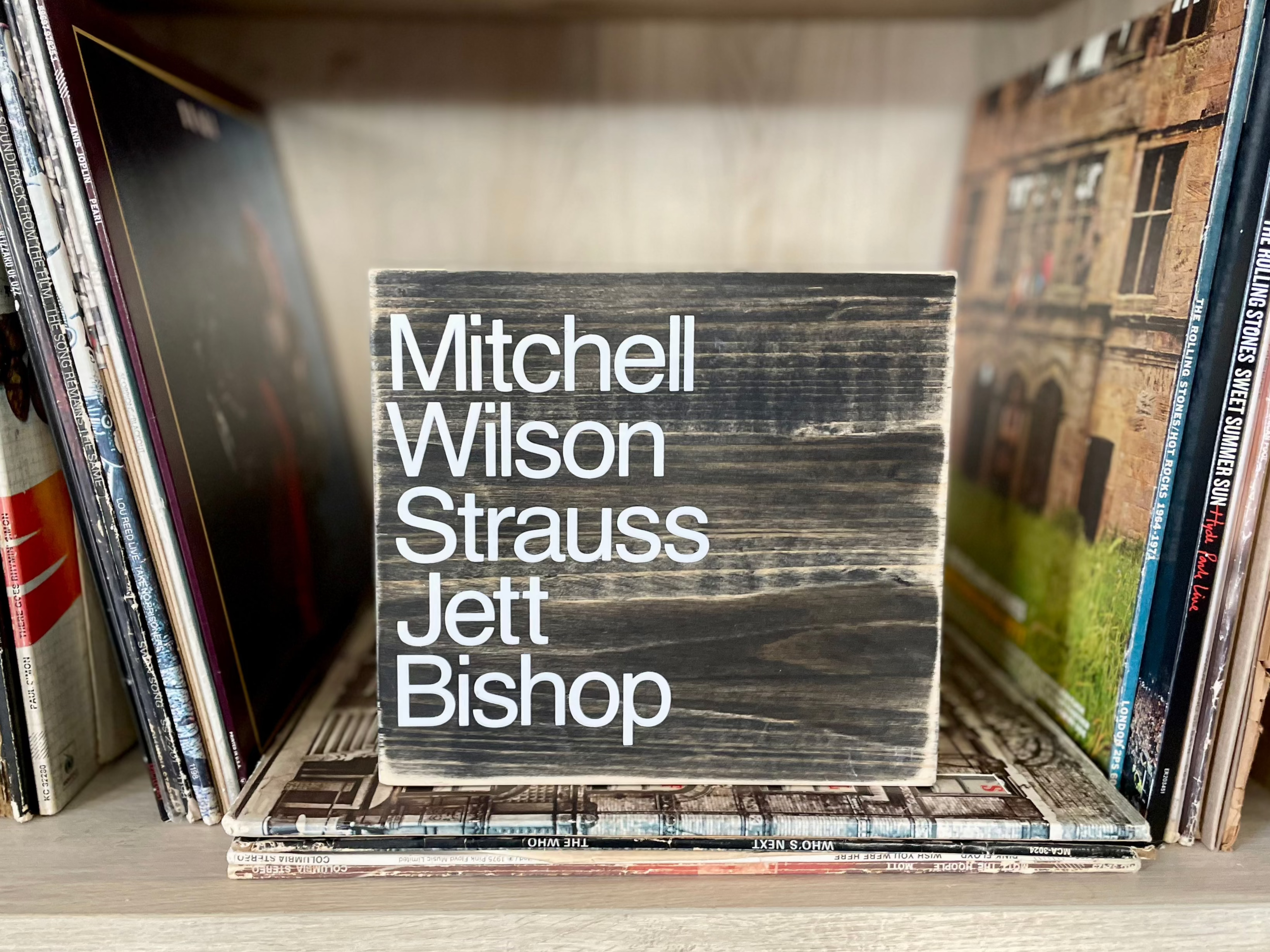 A square, black, wood sign sits on a shelf of  vinyl records 