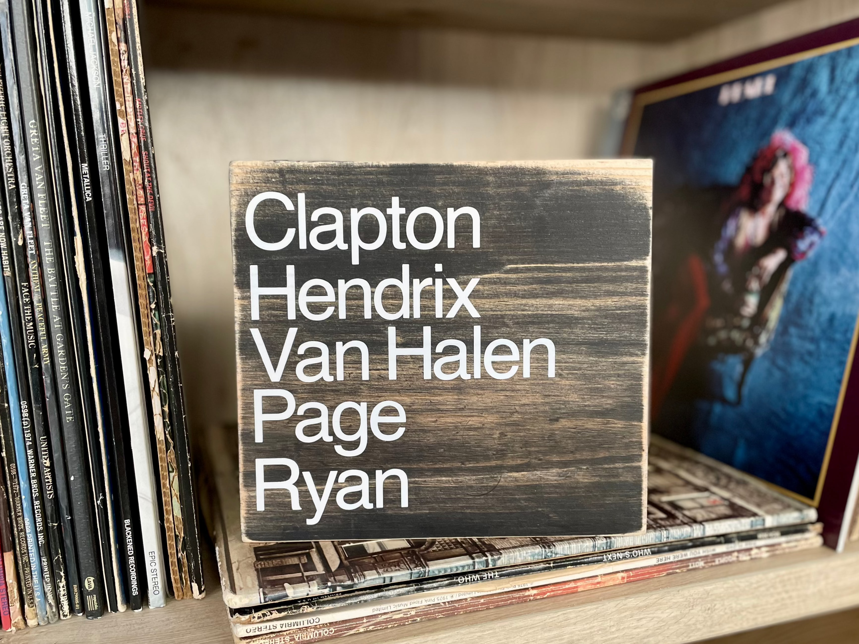 A square, black, wood, sign sits on a shelf with vinyl records.