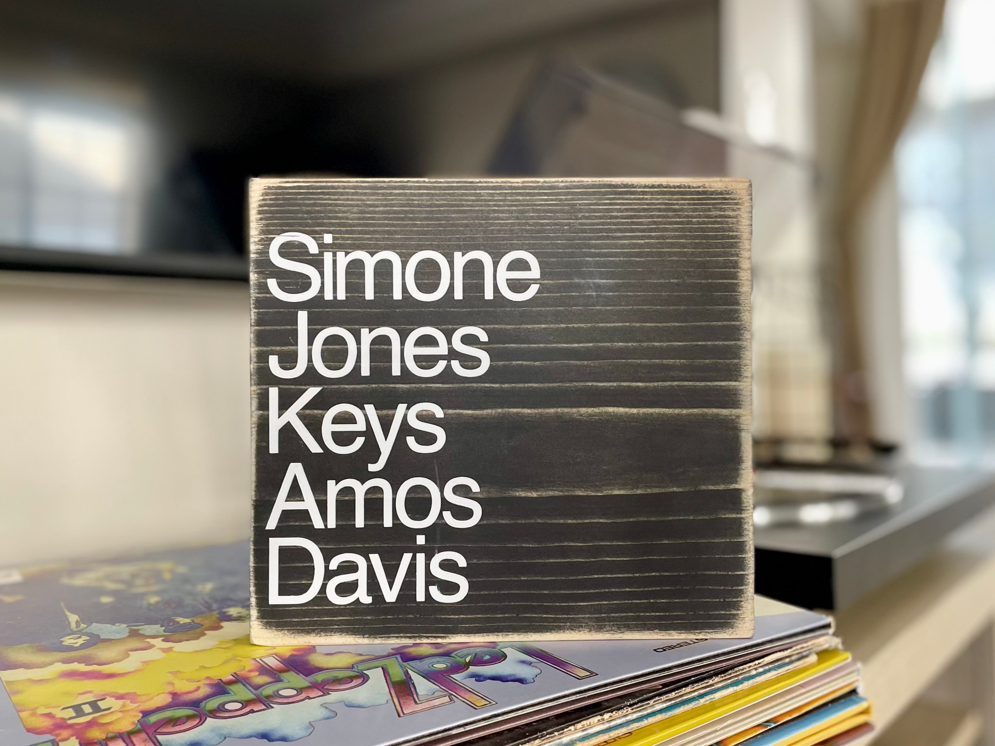 A square, black, wood sign sits on a table next to a record player