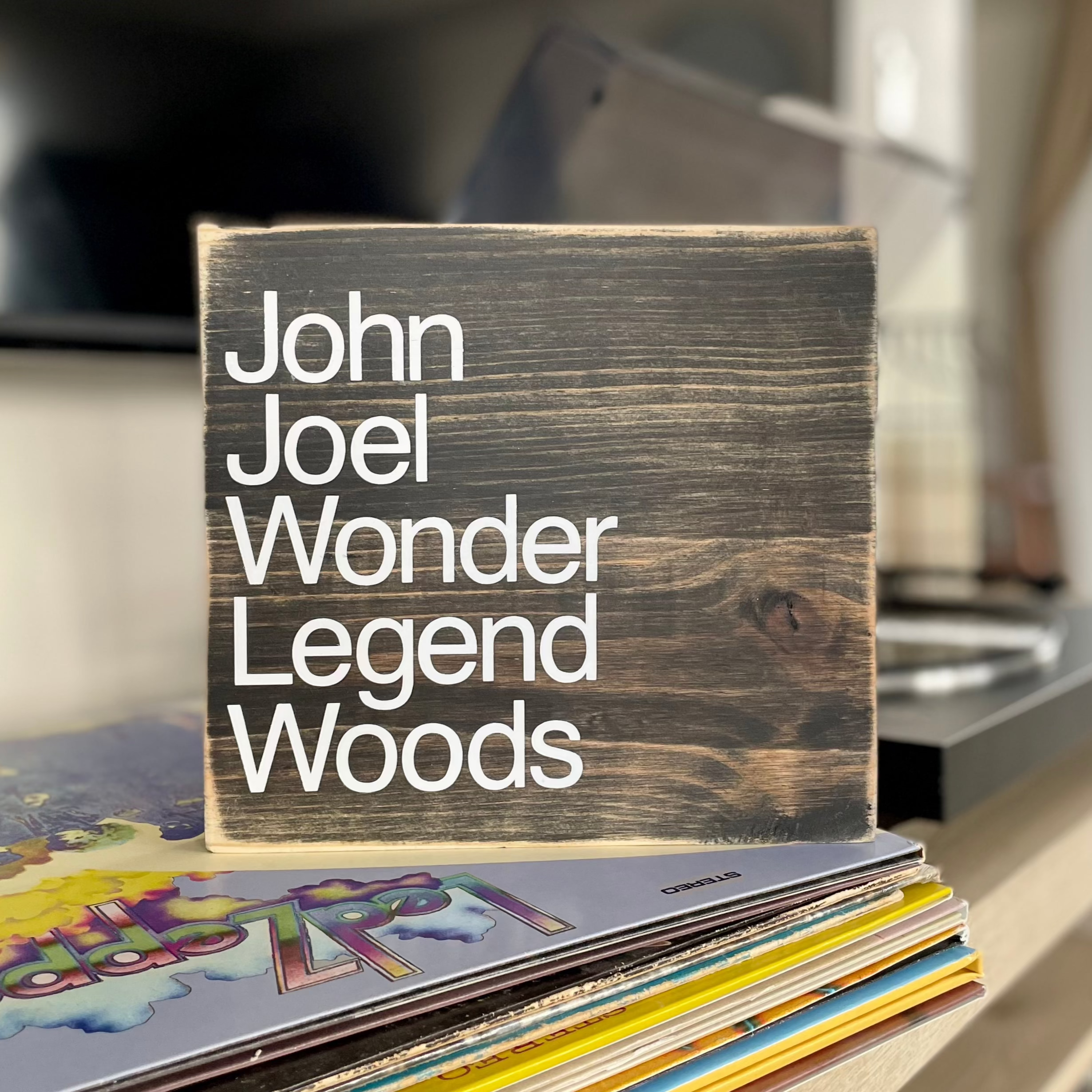 A square, black, wood sign sits on a table next to a record player