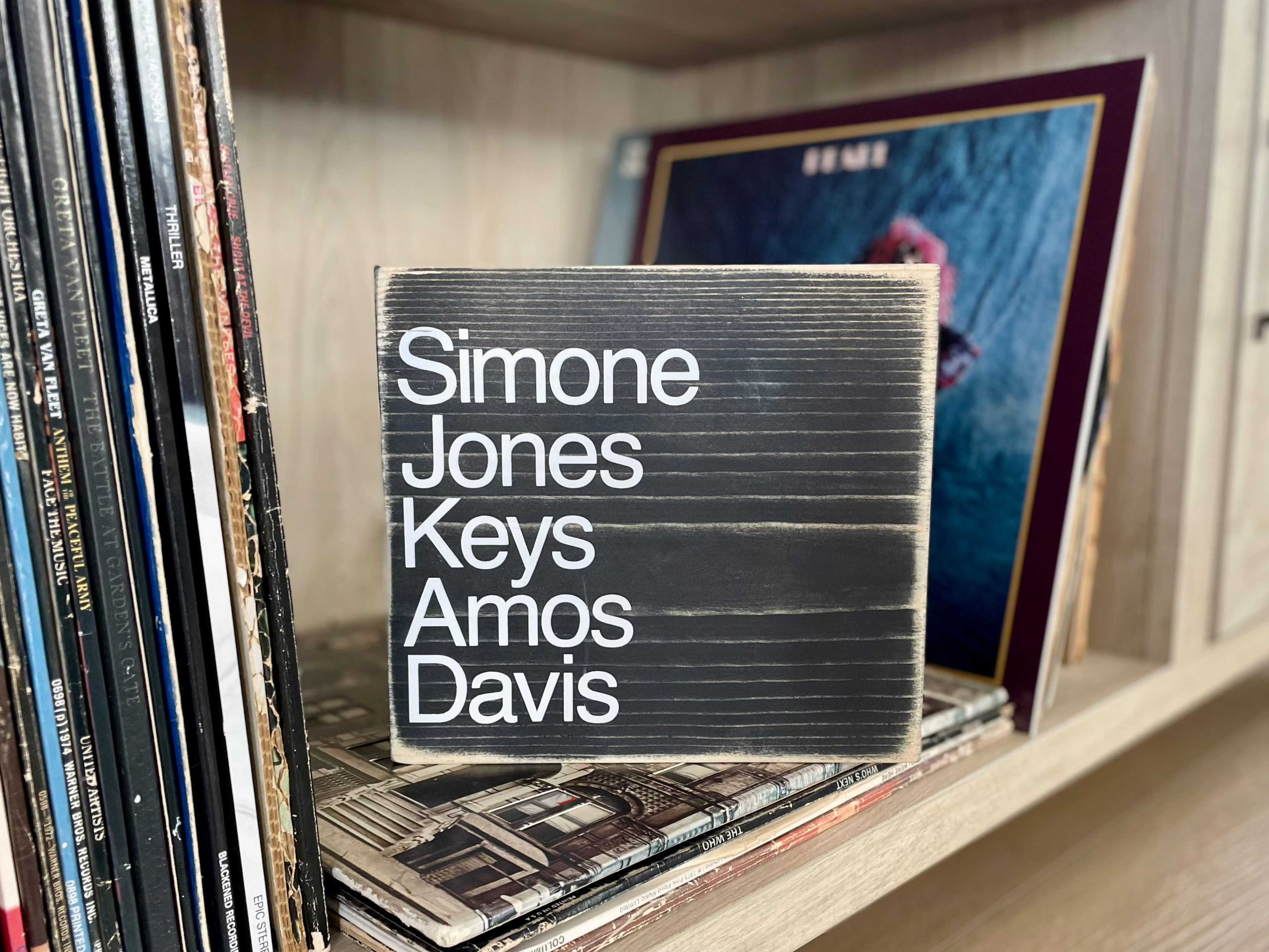 A square, black, wood sign sits on a shelf full of vinyl records 