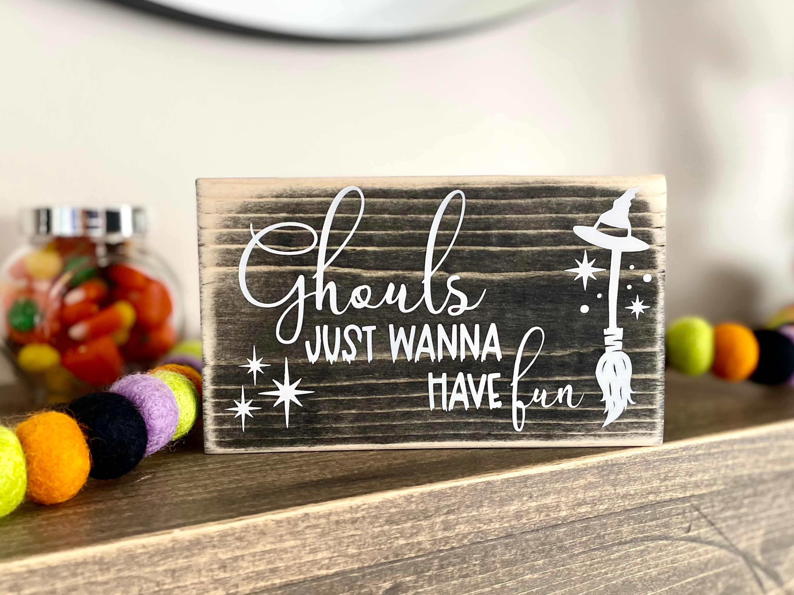 A small, black, wood sign sits on a mantle with Halloween themed decorations behind it.