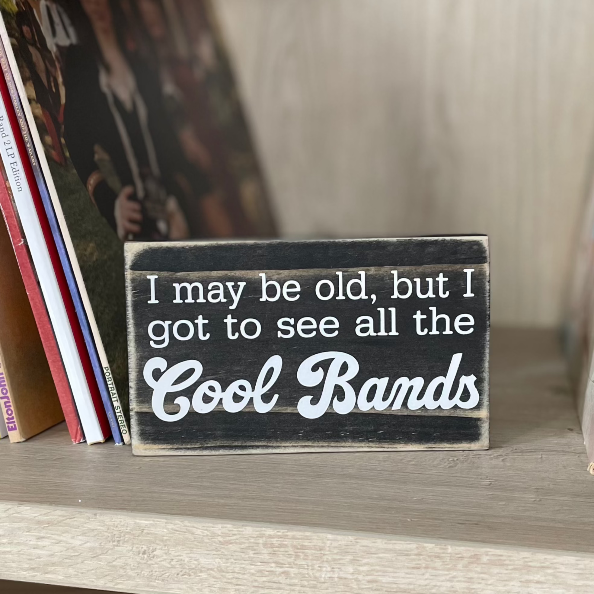 A small, black, wood sign sits on a shelf next to stacks of vinyl records.