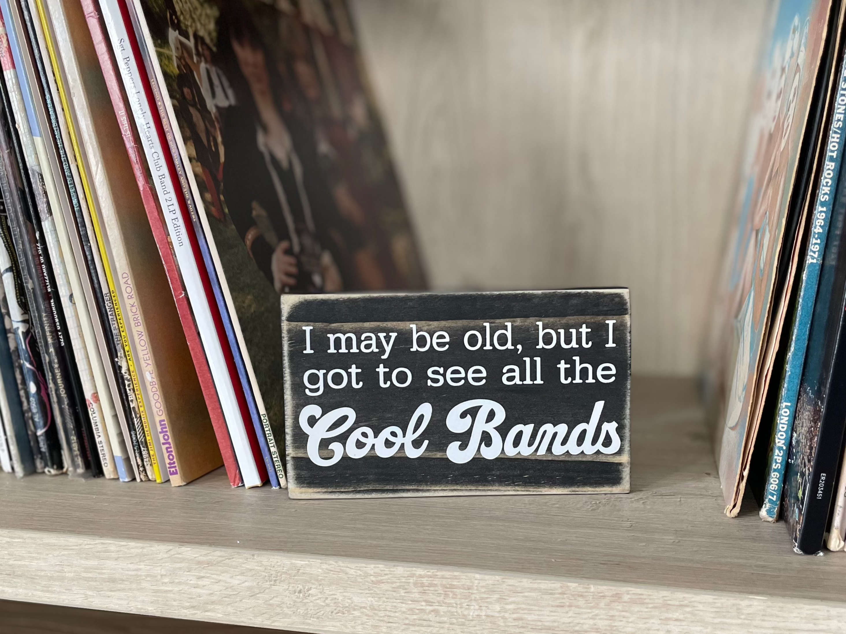 A small, black, wood sign sits on a shelf next to stacks of vinyl records.