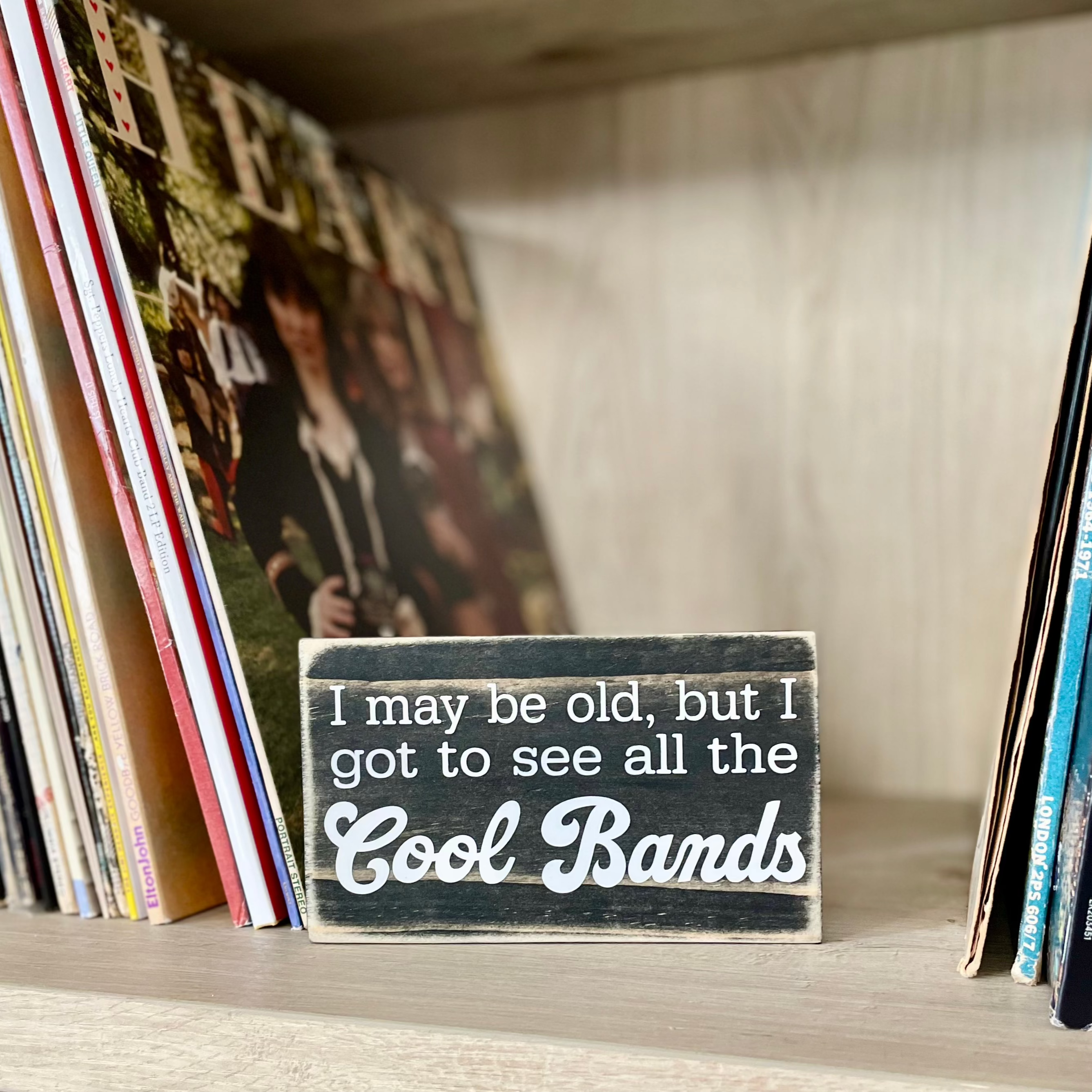 A small, black, wood sign sits on a shelf next to stacks of vinyl records.