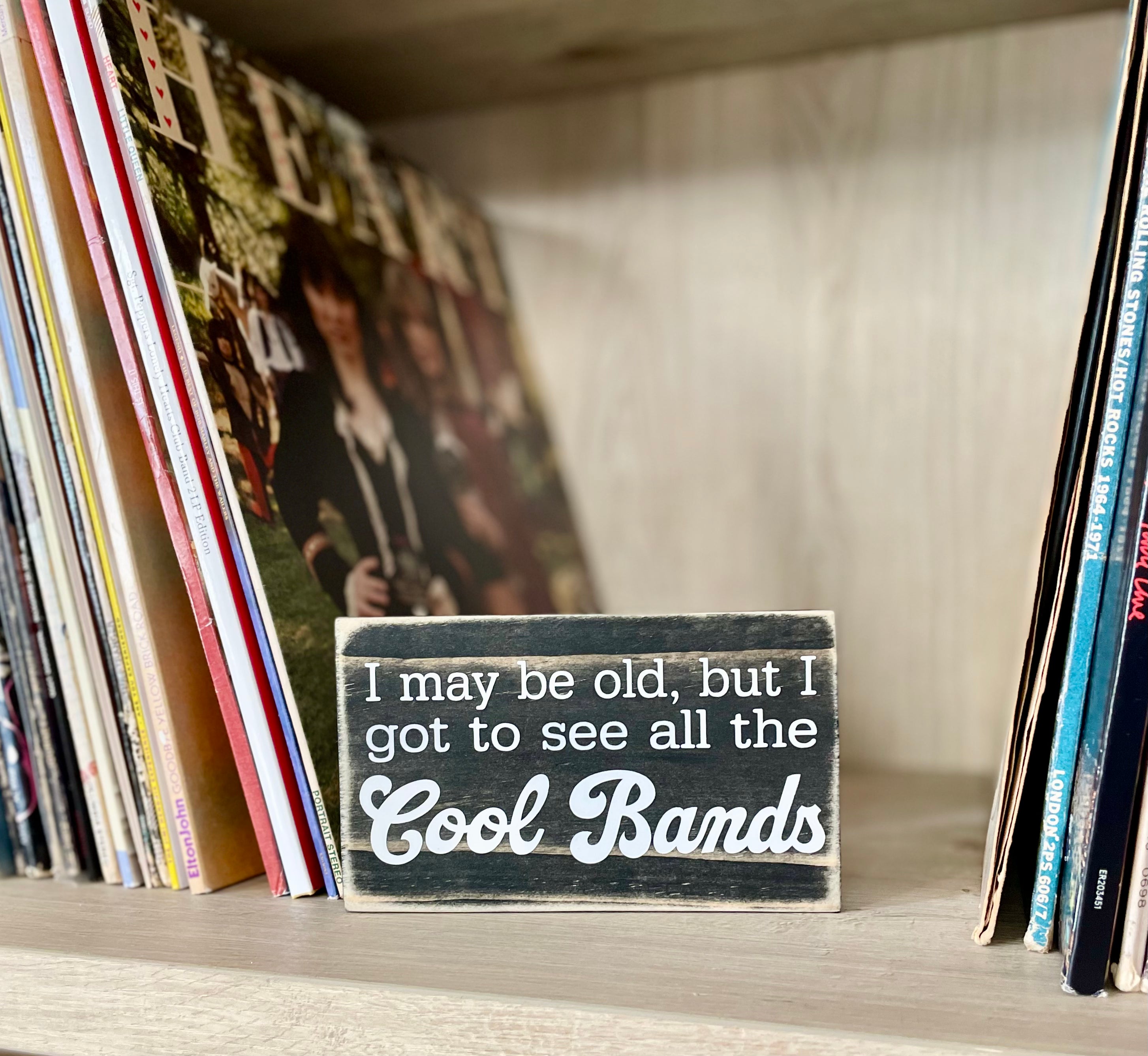 A small, black, wood sign sits on a shelf next to stacks of vinyl records.