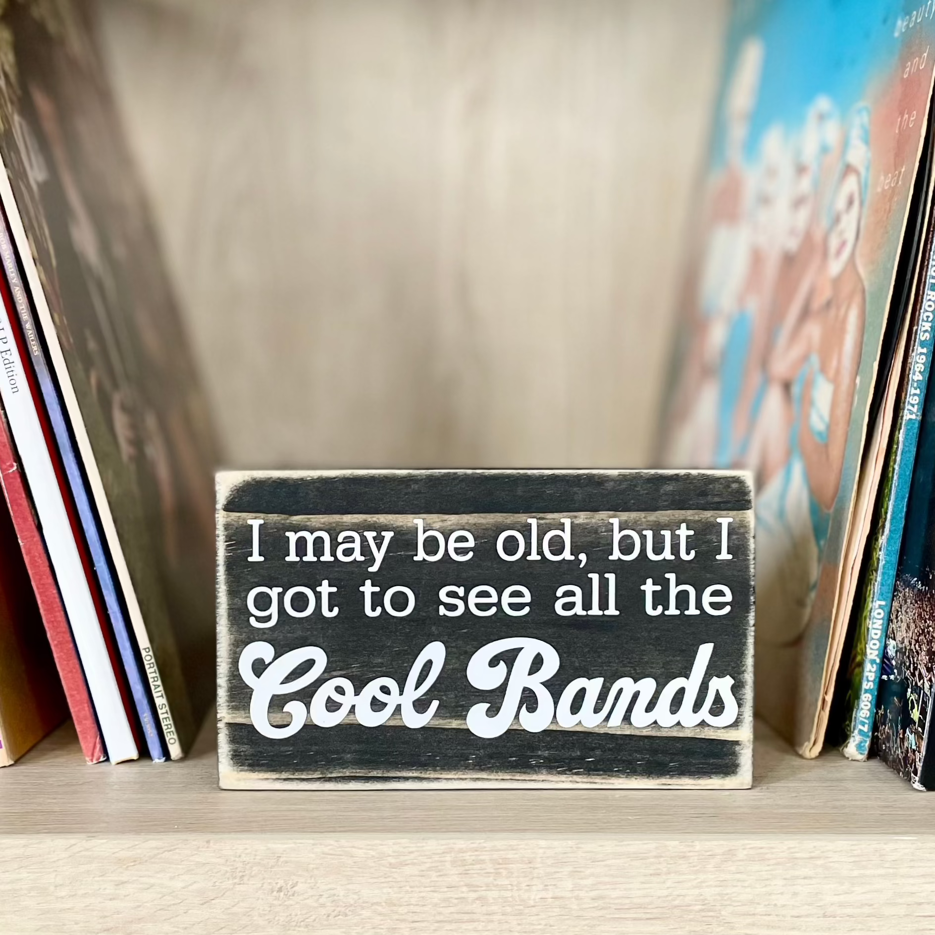 A small, black, wood sign sits on a shelf next to stacks of vinyl records.