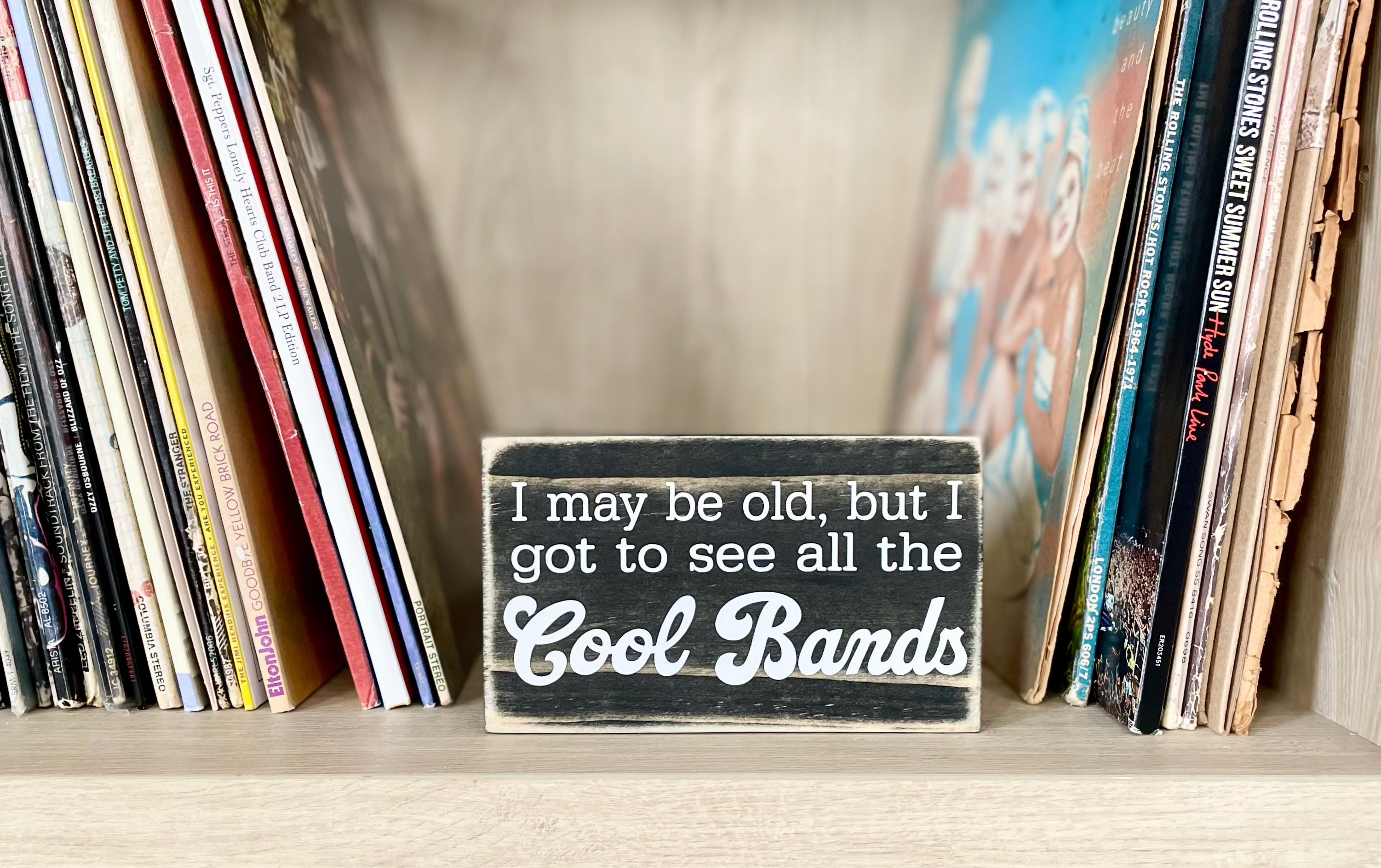 A small, black, wood sign sits on a shelf next to stacks of vinyl records.