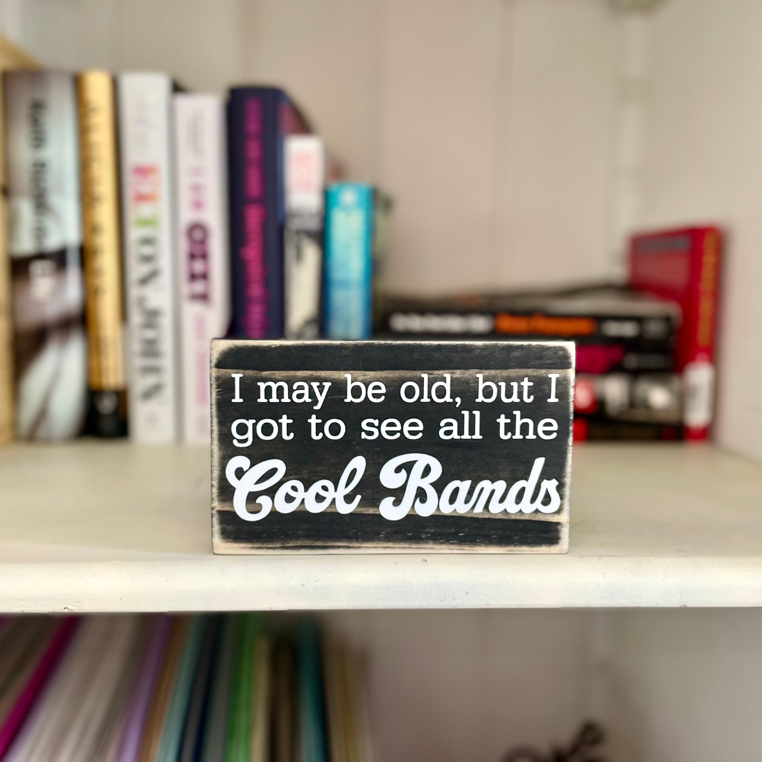 A small, black, wood sign sits on a shelf full of rock biography books.