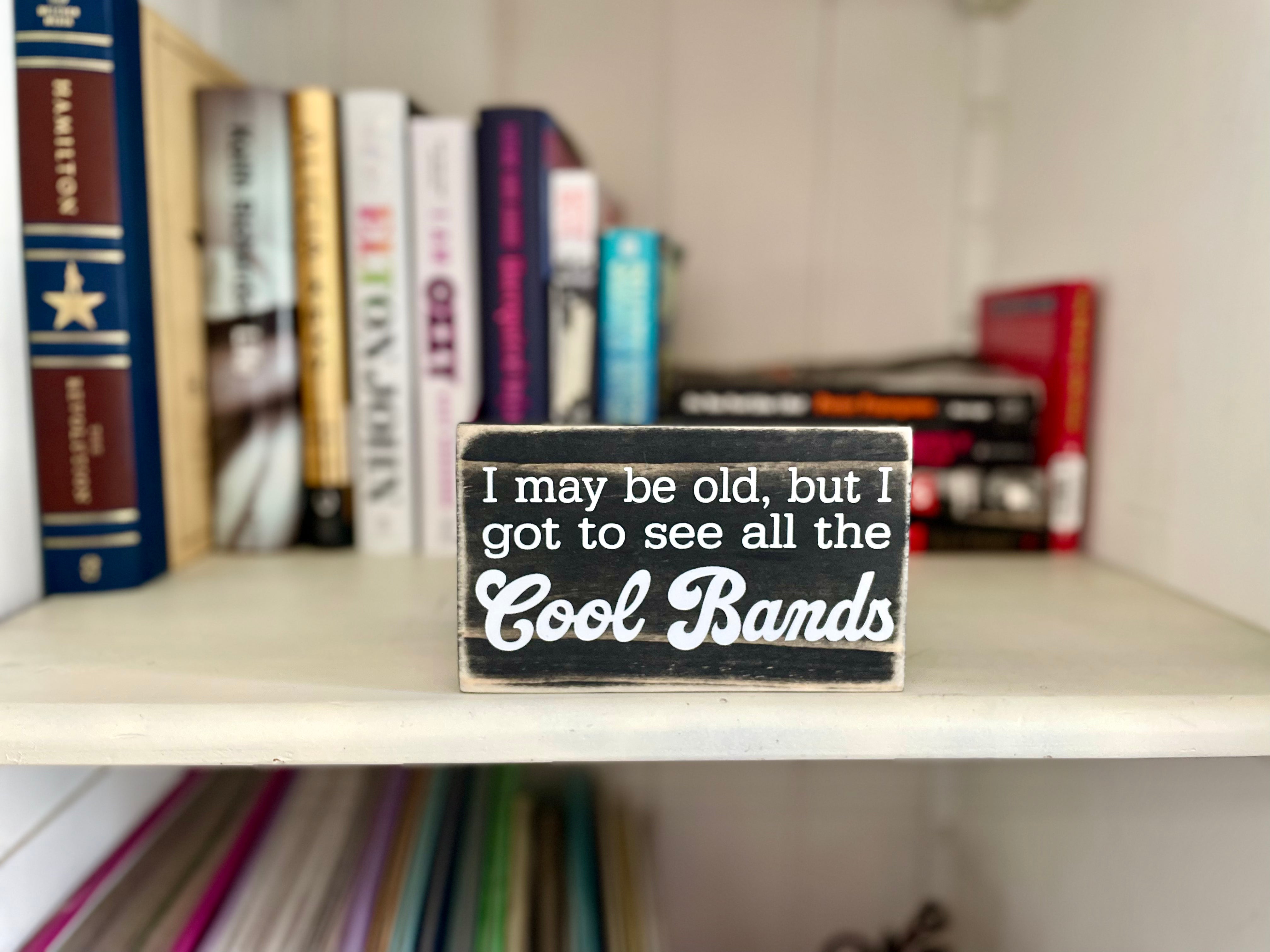 A small, black, wood sign sits on a shelf full of rock biography books.