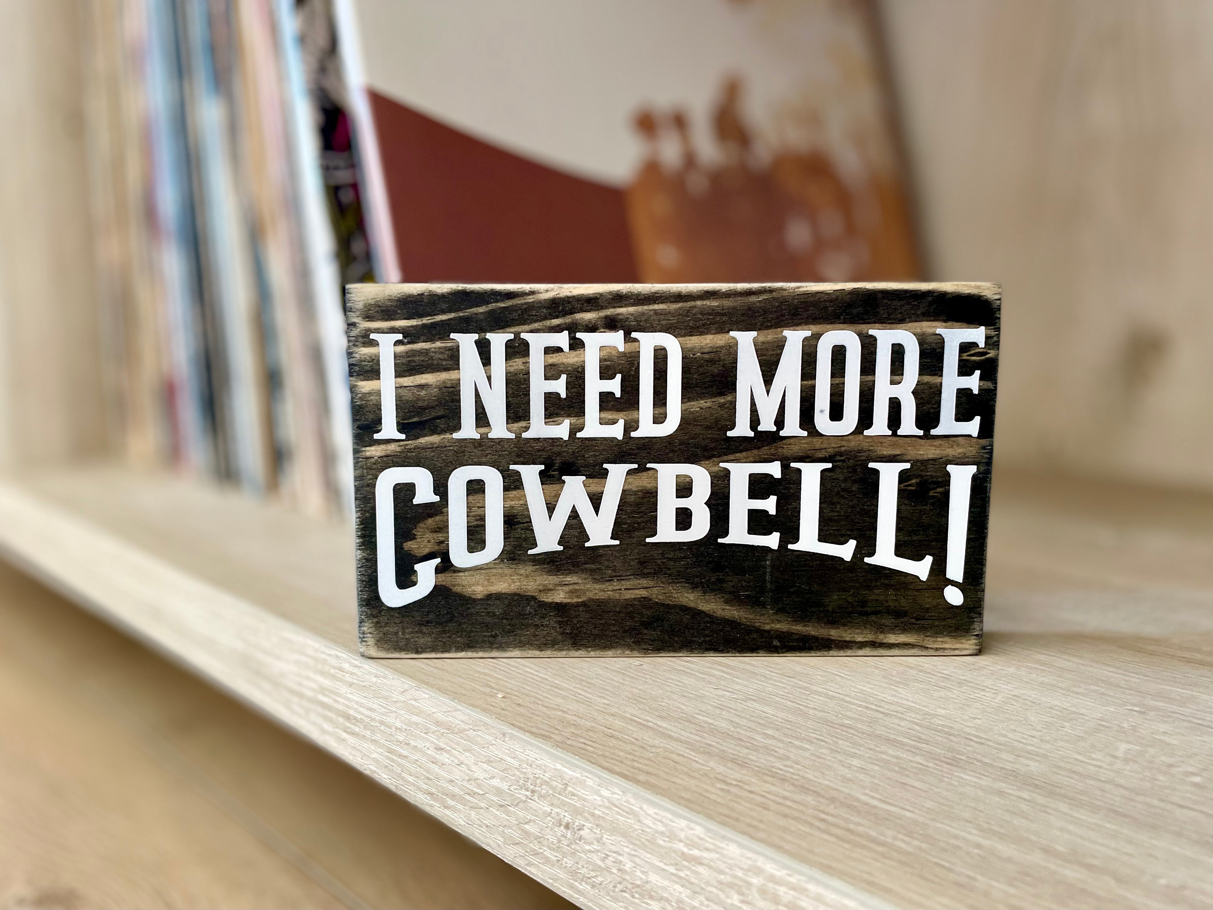 A small, black, wood sign sits on a bookshelf with stacks of vinyl records blurred in the background. 