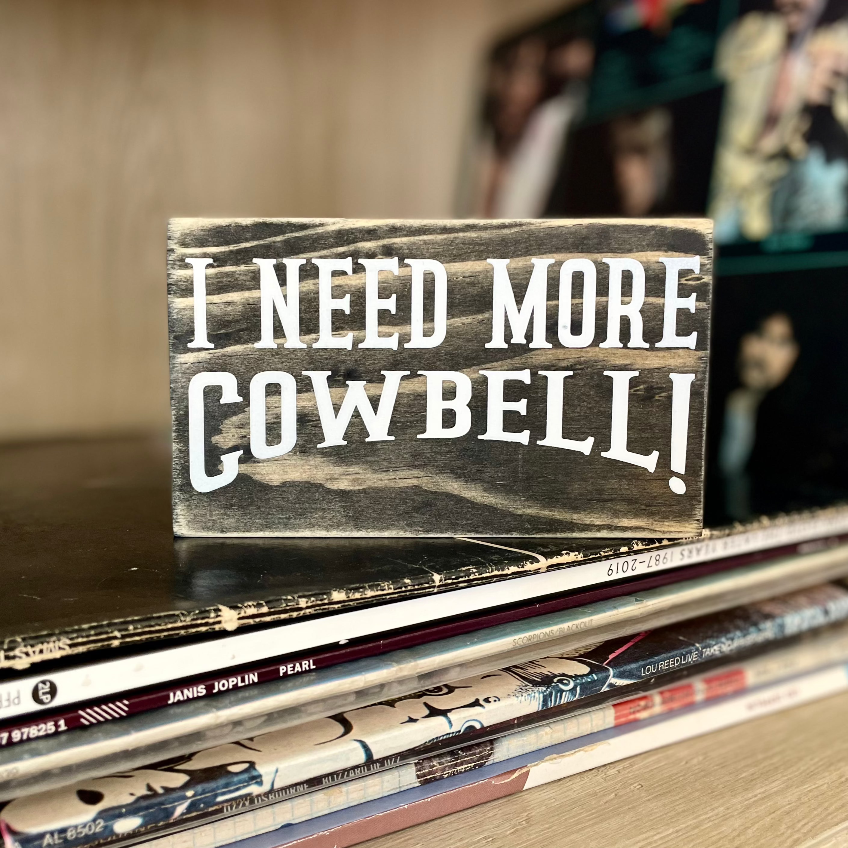 A small, black, wood sign sits on a stack of vinyl records with more records blurred in the background. 