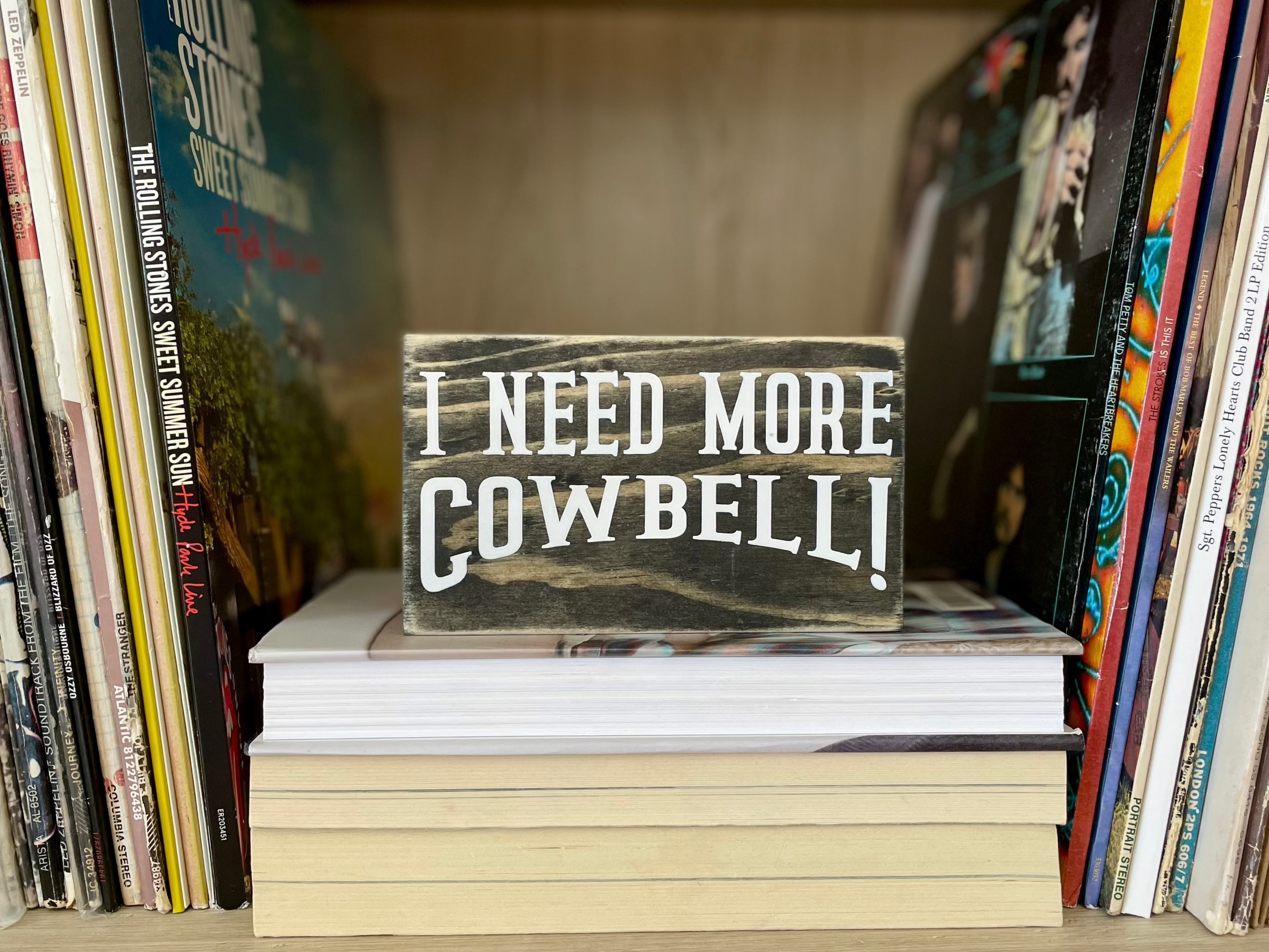 A small, black, wood sign sits on stack of books with stacks of vinyl records on both sides of it.