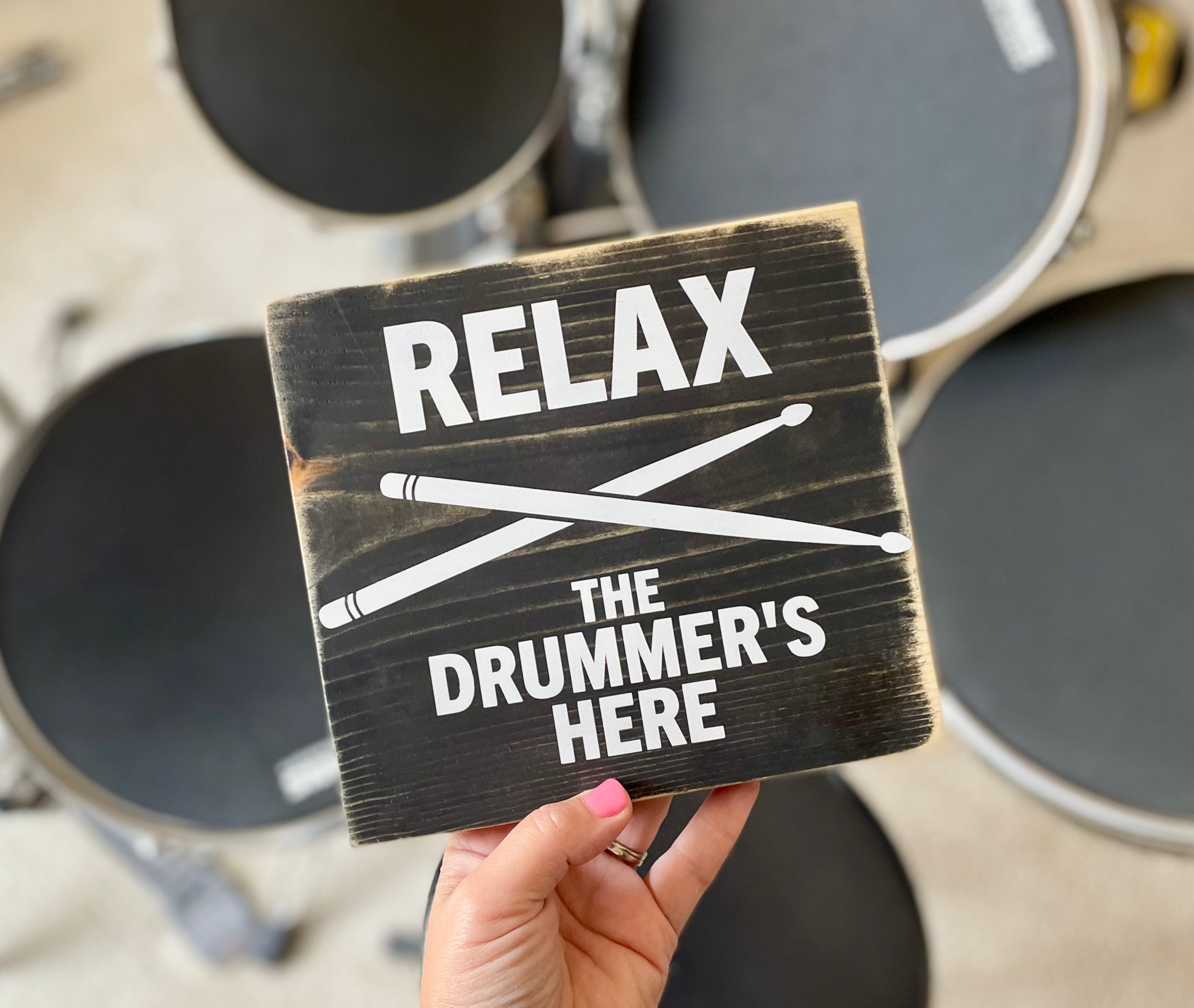 A black, wood sign with white lettering is being held in front of a drum kit that is blurred in the background. The sign reads: "Relax the Drummer's Here." A white pair of drumsticks is painted in an x under the word relax.